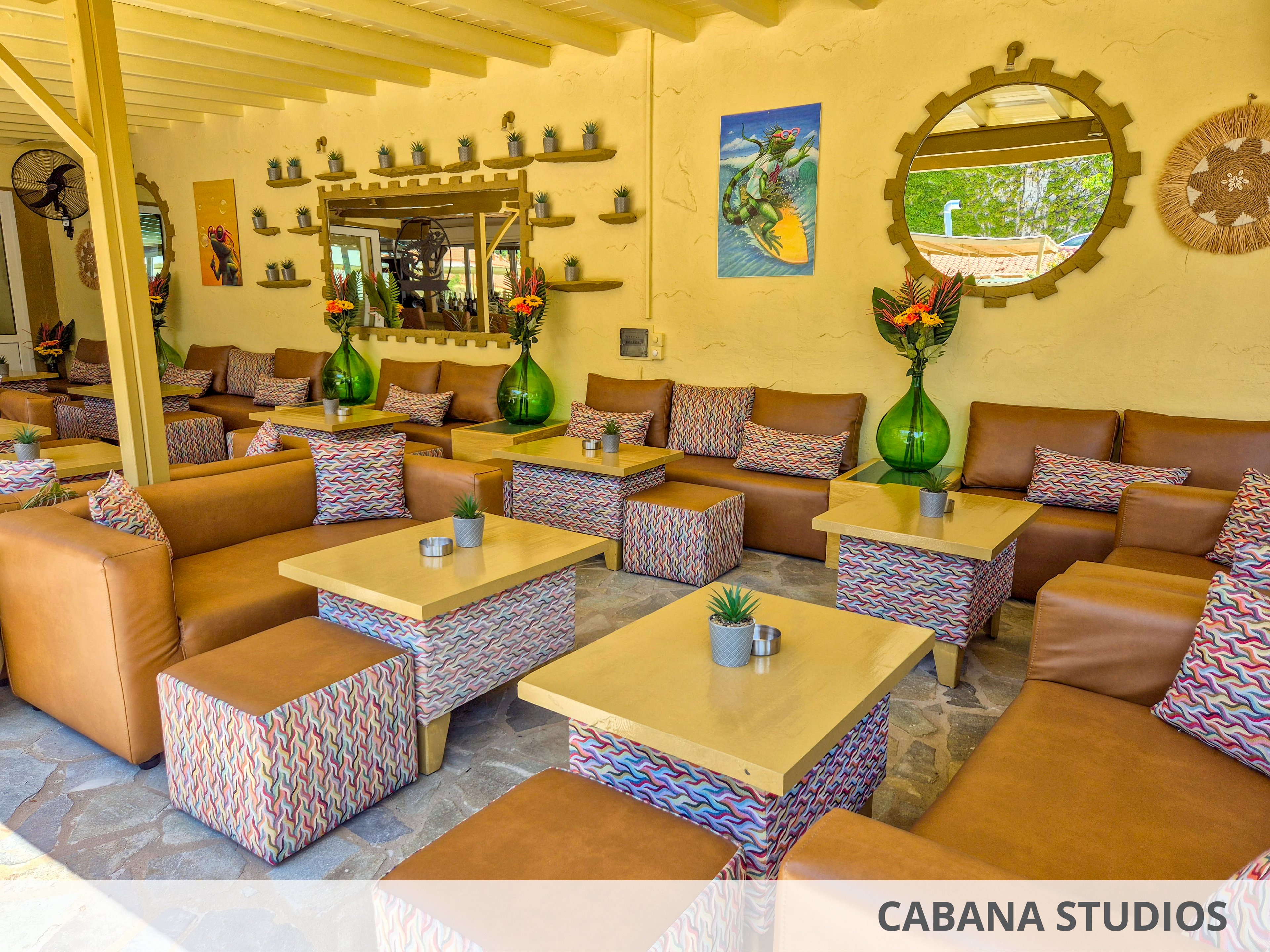 A bright outdoor lounge area features multiple tan leather sofas and matching cube ottomans arranged around light wooden tables. Patterned fabric on the ottomans and cushions adds colorful geometric accents to the space. Large green glass vases with tropical flower arrangements decorate the tabletops, while small potted succulents provide additional greenery. The background wall is adorned with mirrors, shelves of small pots, and vibrant artwork, creating a relaxed yet stylish social setting.