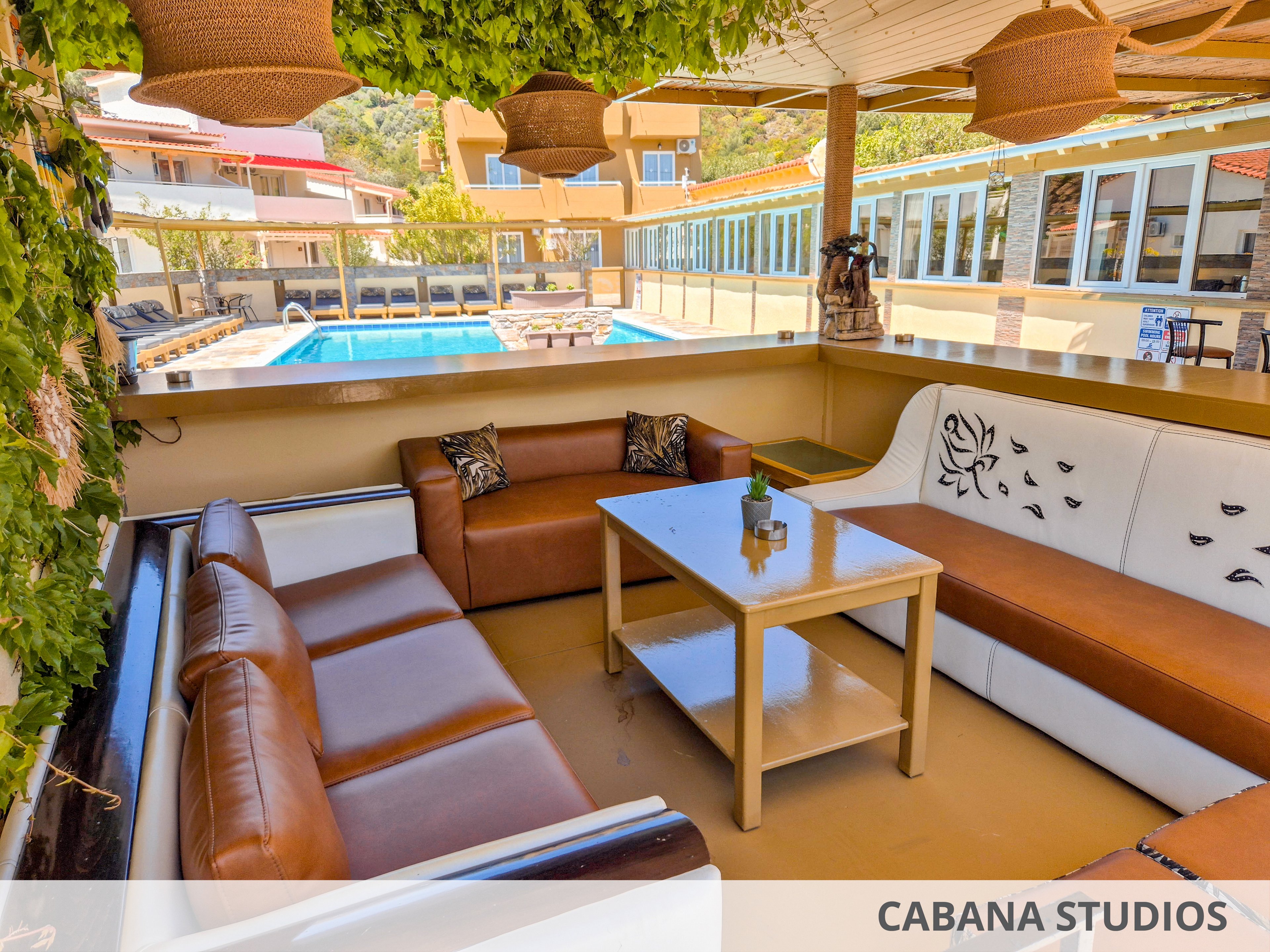 An outdoor lounge area features brown and white cushioned sofas arranged around a rectangular coffee table. The seating space is shaded by a pergola with hanging woven light fixtures and green vines. In the background, a clear blue swimming pool is surrounded by buildings with large windows and colorful roofs. The scene suggests a relaxed, resort-like environment ideal for socializing and leisure.