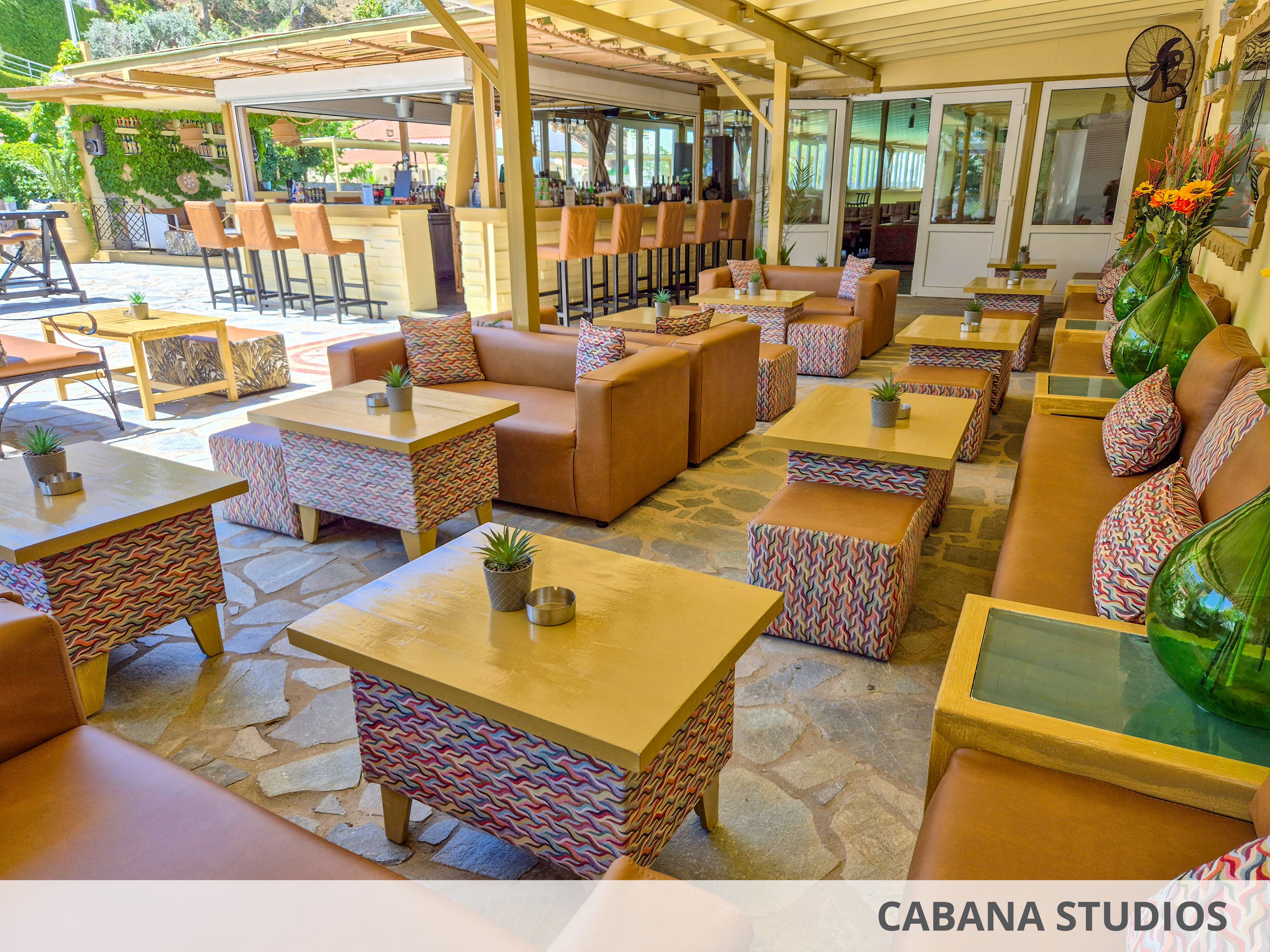 An outdoor lounge area features tan leather sofas and patterned cube ottomans arranged around low wooden tables. The space opens onto a bar with high stools under a covered pergola roof. Small potted plants and candles decorate the tables, adding a relaxed and inviting touch. Large glass vases with flowers and greenery accent the seating area, while natural stone flooring enhances the casual resort atmosphere.