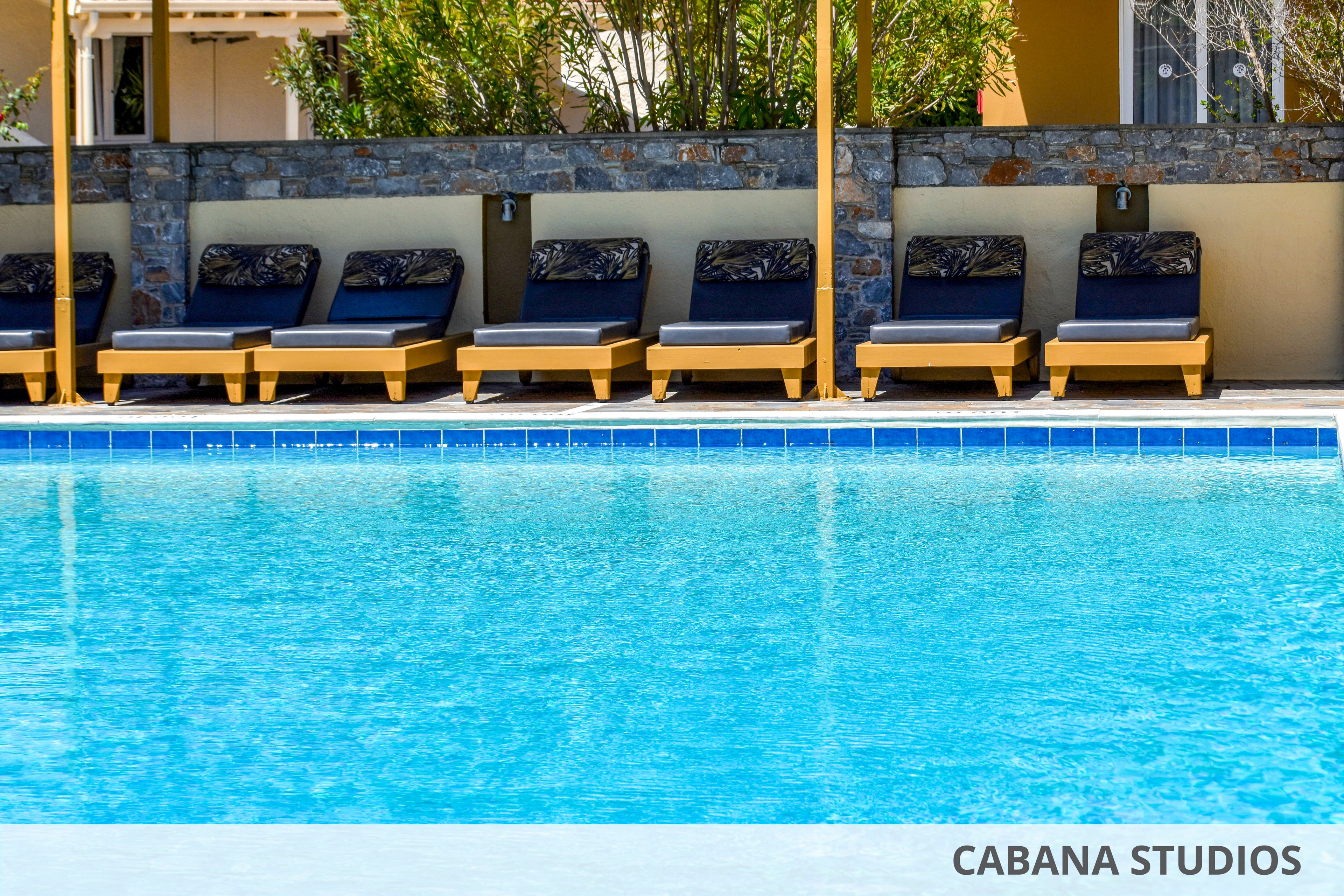 A clear blue swimming pool fills the foreground of the image. Behind the pool, a row of cushioned sun loungers sits neatly aligned under partial shade. A low stone wall and green shrubs create a private, relaxed backdrop. The scene suggests a quiet resort or hotel area ready for guests to relax by the water.