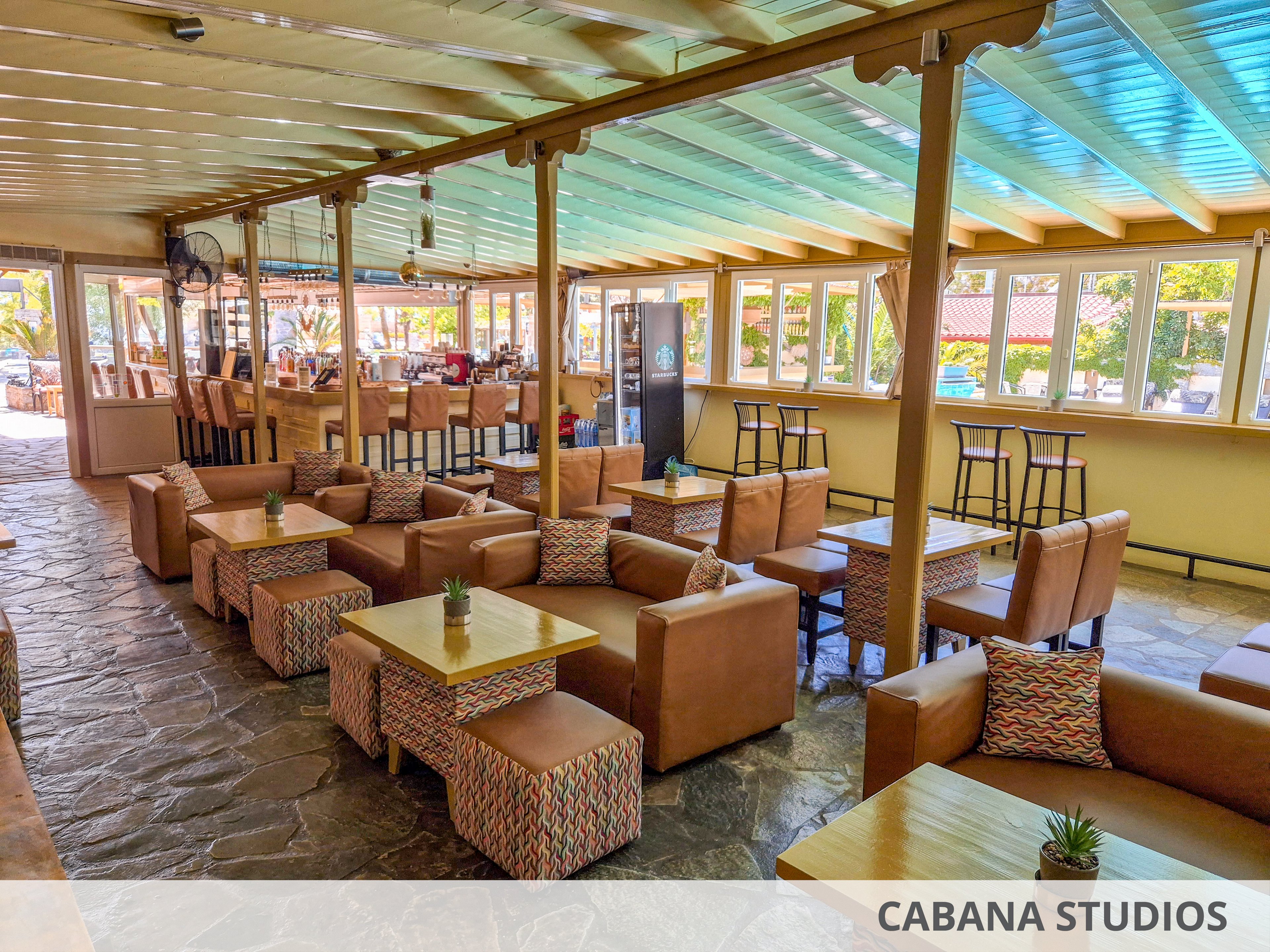 Spacious indoor lounge bar with rows of cushioned armchairs and low wooden tables. Large windows line the wall, letting in bright daylight and views of greenery outside. A bar counter with high stools is visible in the background, along with additional tall tables near the windows. Stone flooring, patterned pillows, and small potted plants on the tables create a relaxed, casual atmosphere.
