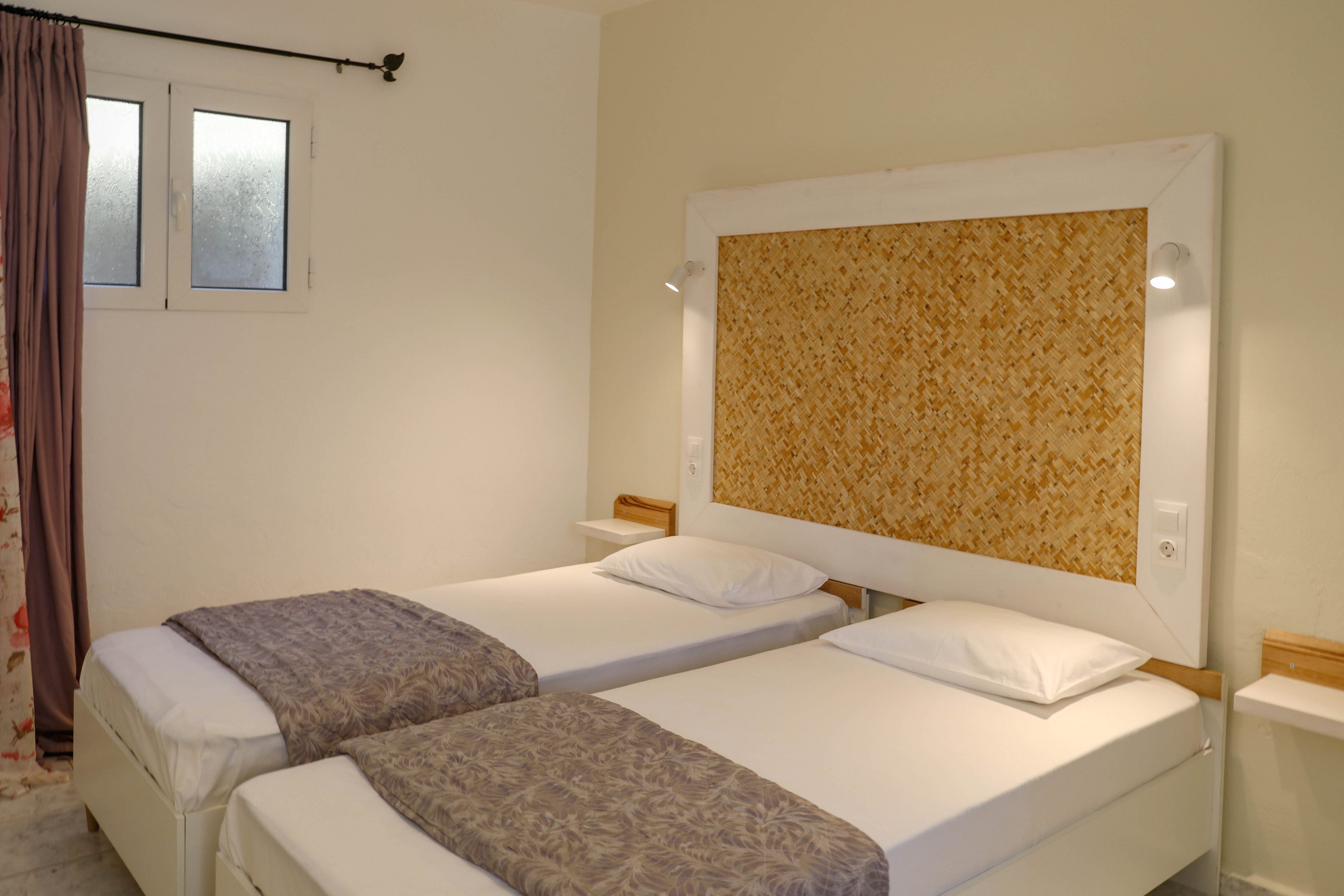A small bedroom features two neatly made twin beds with white linens and patterned gray throws. The beds sit side by side beneath a large textured headboard with built-in reading lights. A curtained window lets in soft natural light, complementing the warm neutral walls. The overall atmosphere is simple, tidy, and inviting, suggesting a comfortable hotel or guest room stay.
