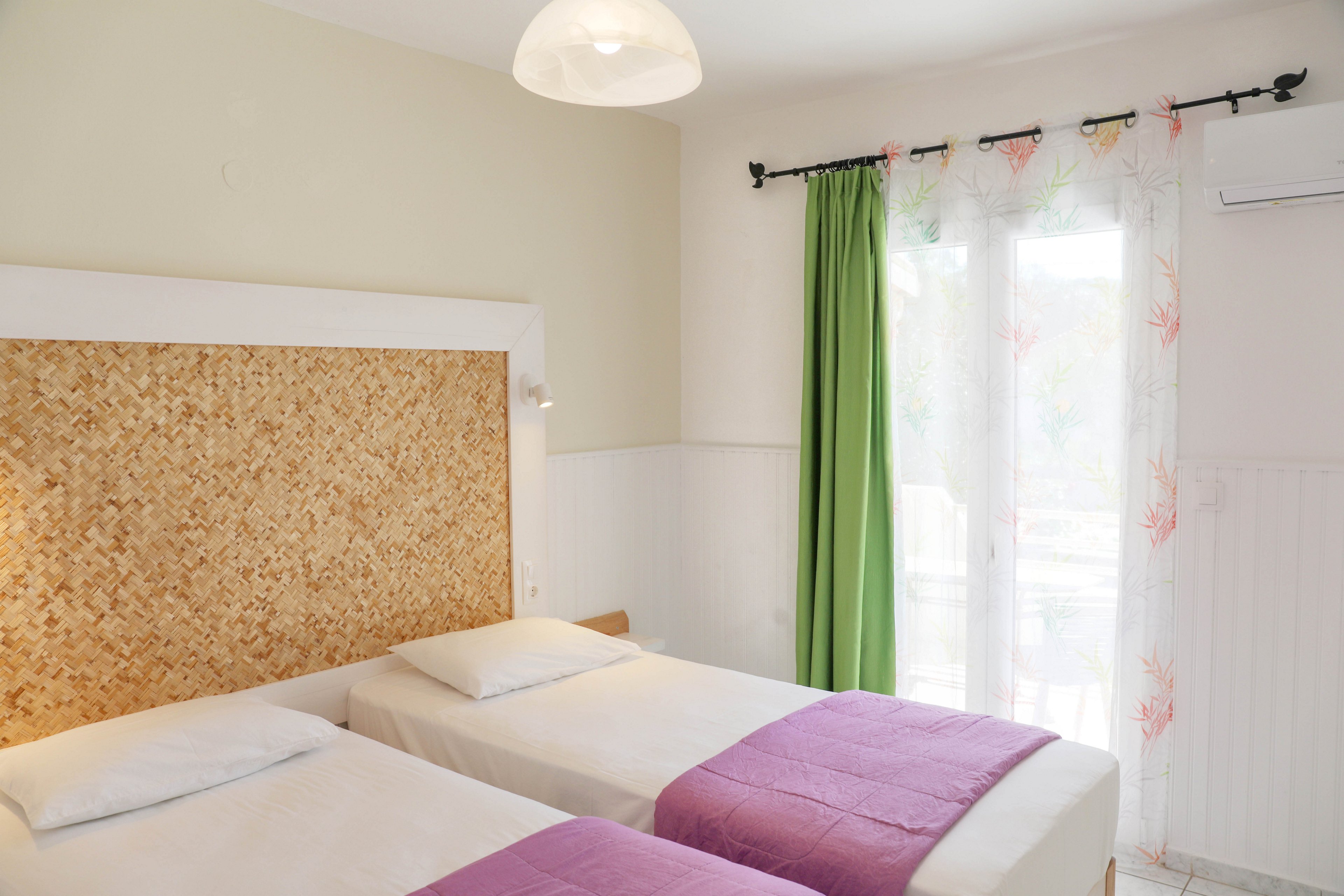A bright bedroom features two twin beds with white linens and purple throws. The beds share a large woven headboard that adds texture to the minimalist decor. Sheer curtains with a subtle pattern hang beside a vivid green drape over a glass door, allowing natural light to fill the room. A simple ceiling lamp and white walls contribute to the clean, airy atmosphere.