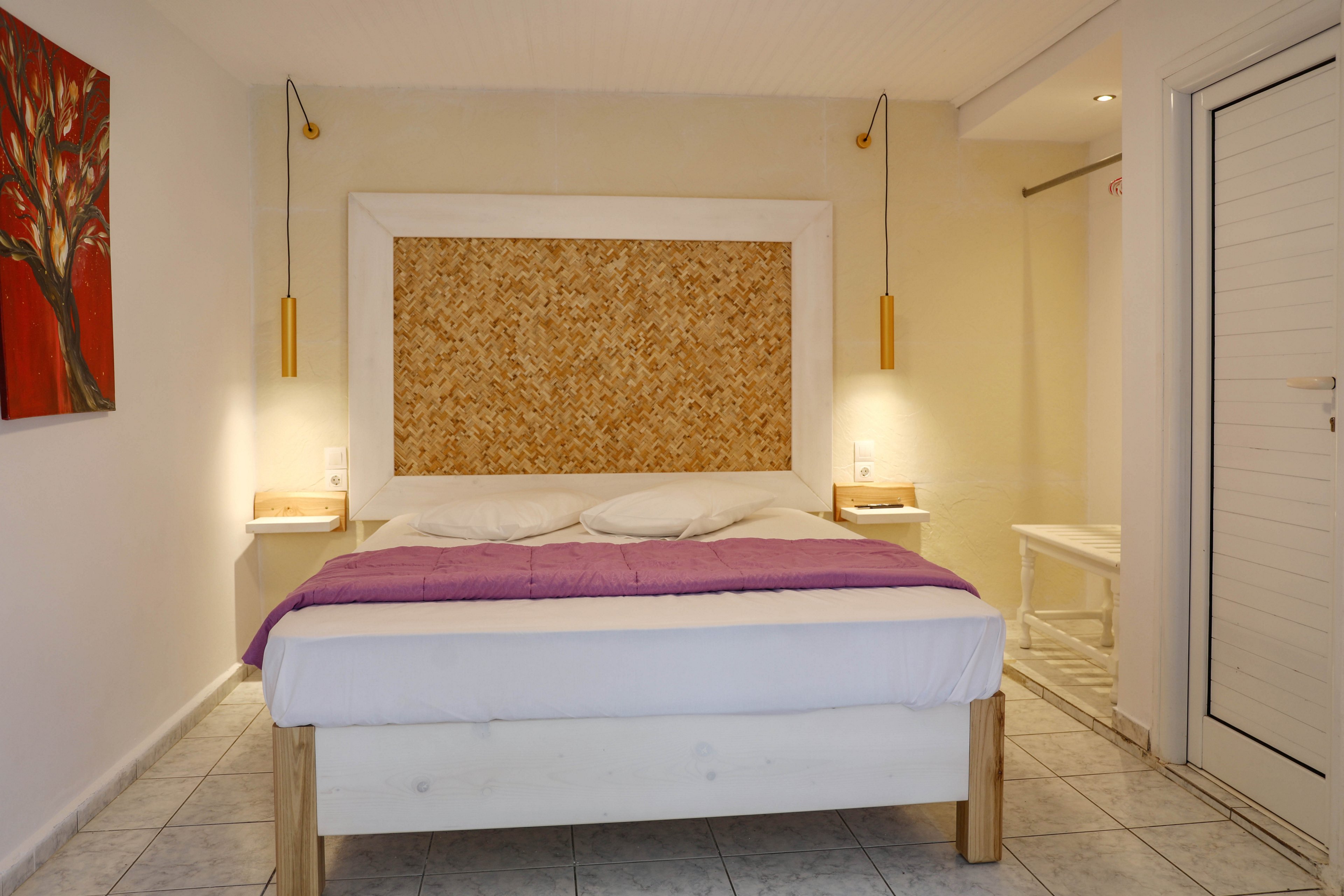 A neatly made double bed with a purple blanket stands against a textured headboard in a minimalist bedroom. Warm pendant lights hang on either side above small floating bedside tables. The room features light tiled flooring, white walls, and a simple door to the right. A colorful tree painting adds a touch of art and contrast to the otherwise neutral decor.