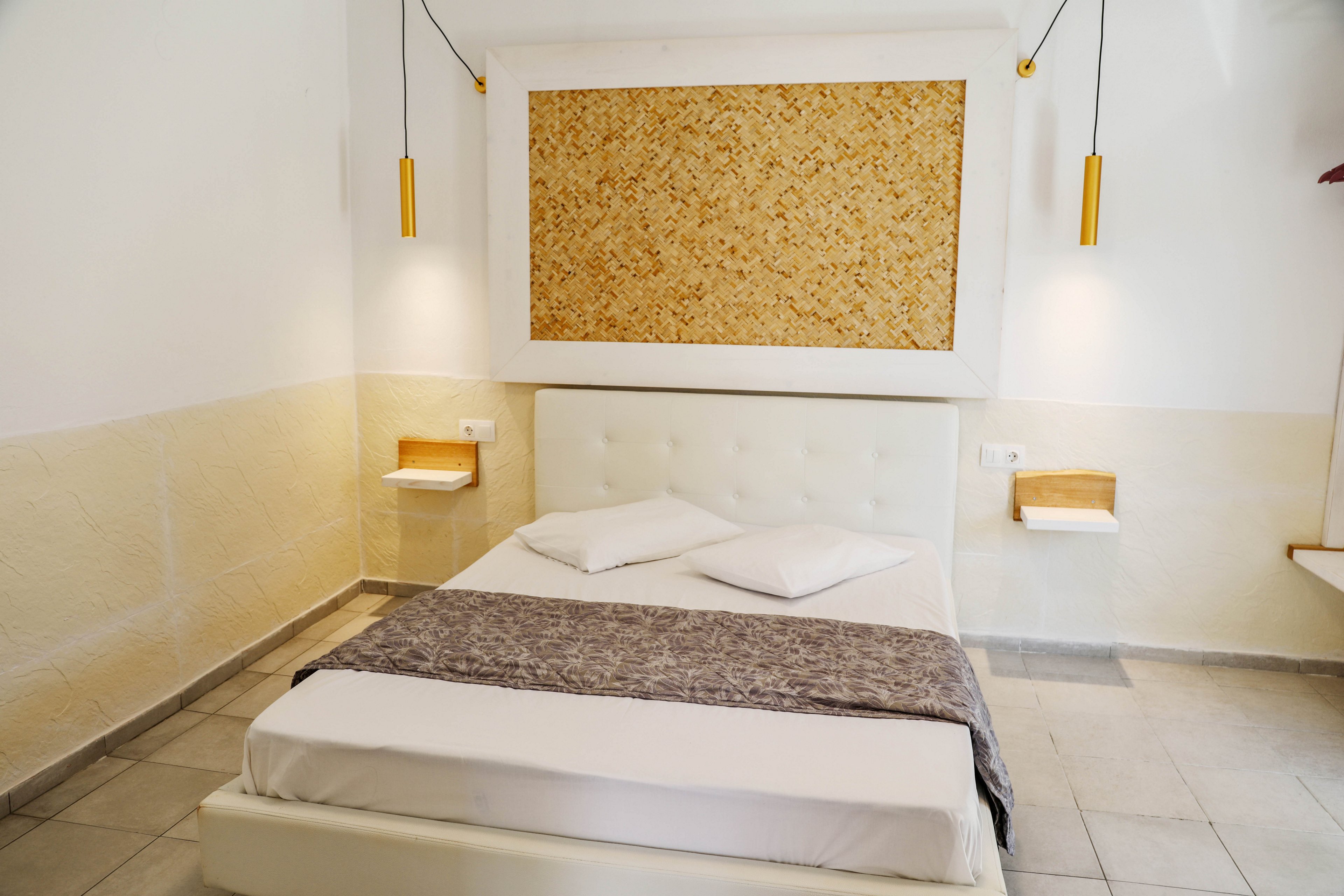 A simple white bed with a tufted headboard is placed against a light-colored wall in a minimalist bedroom. Two pillows and a folded gray throw blanket rest neatly on the bed. Floating bedside shelves with small wooden surfaces flank the bed on both sides. Slim yellow pendant lights hang above the shelves, and a large textured panel decorates the wall above the headboard. The room features neutral tones and clean lines, creating a calm and modern atmosphere.