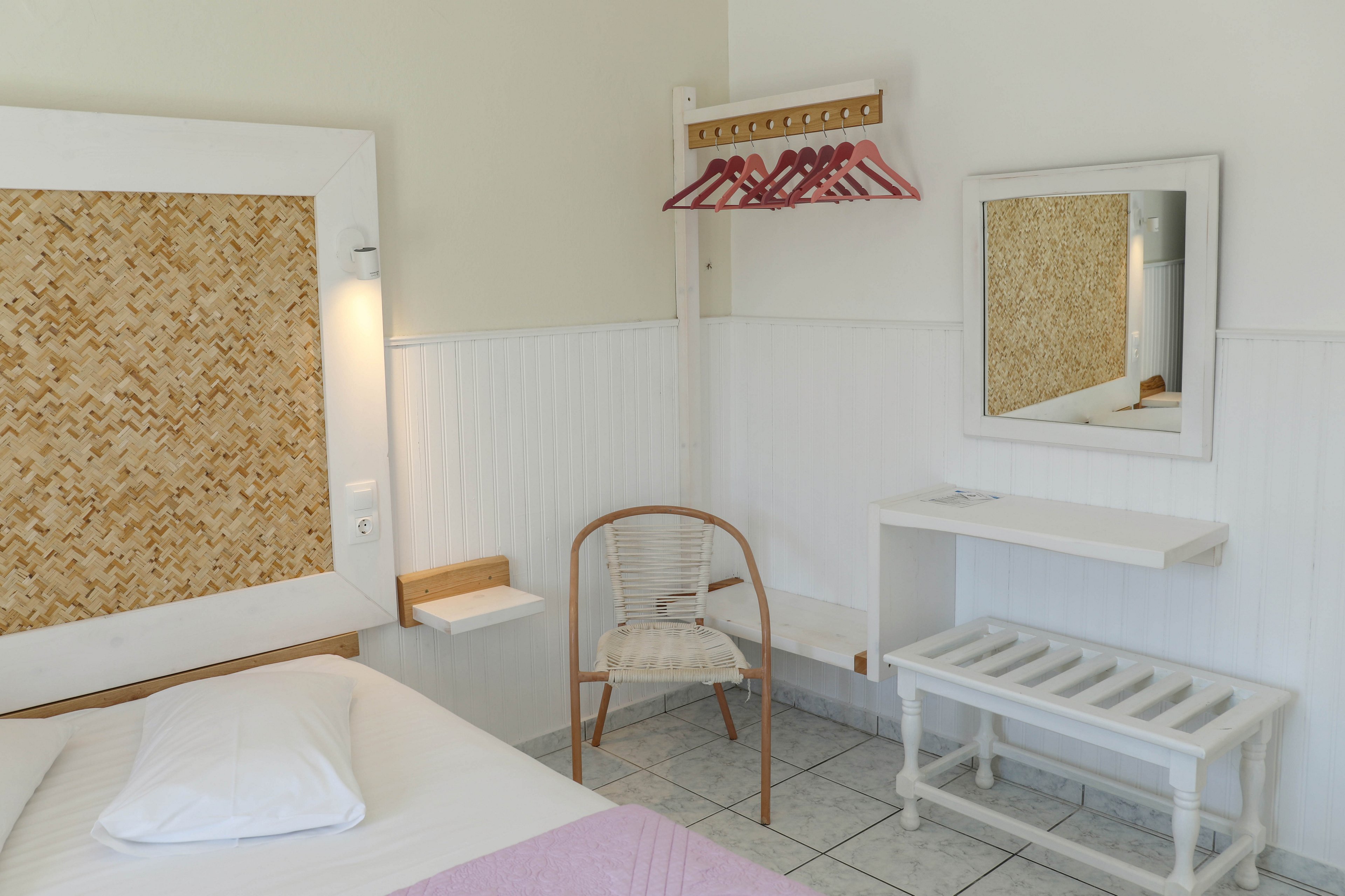 A small, minimalist hotel room features a single bed with a white pillow and light blanket. The space includes a wicker headboard, a simple wooden chair, and a wall-mounted shelf with several red clothes hangers. A white-framed mirror reflects part of the room, adding depth to the bright interior. Light-colored walls and tiled flooring create a clean, airy atmosphere with subtle natural textures.