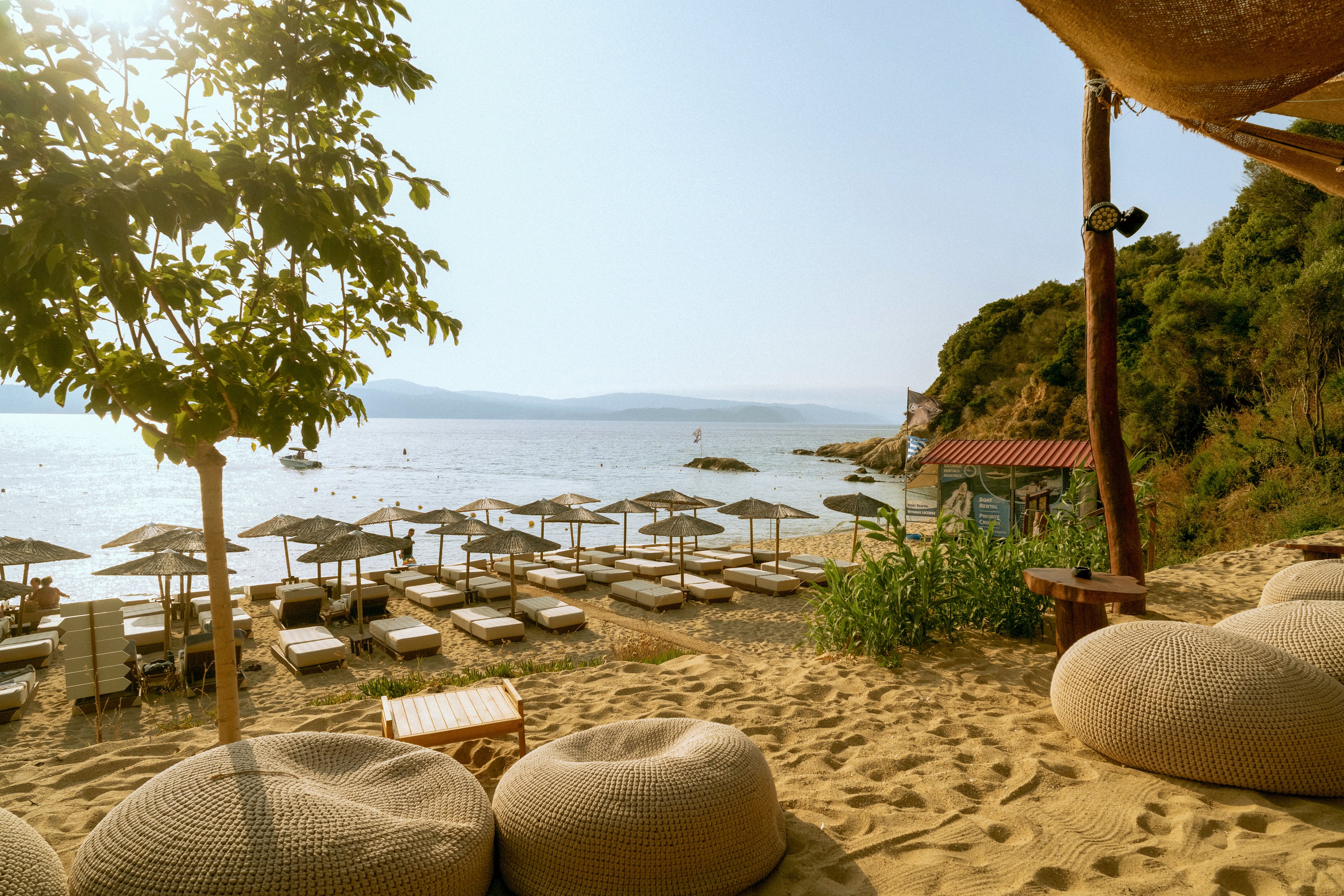 A tranquil beach lounge area is set up with rows of sunbeds and straw umbrellas facing a calm sea. The sandy shore is framed by cozy woven poufs and a shaded seating area in the foreground. Gentle waves and distant swimmers are visible under the warm afternoon light. Green trees and coastal hills border the beach, creating a relaxed summer atmosphere.