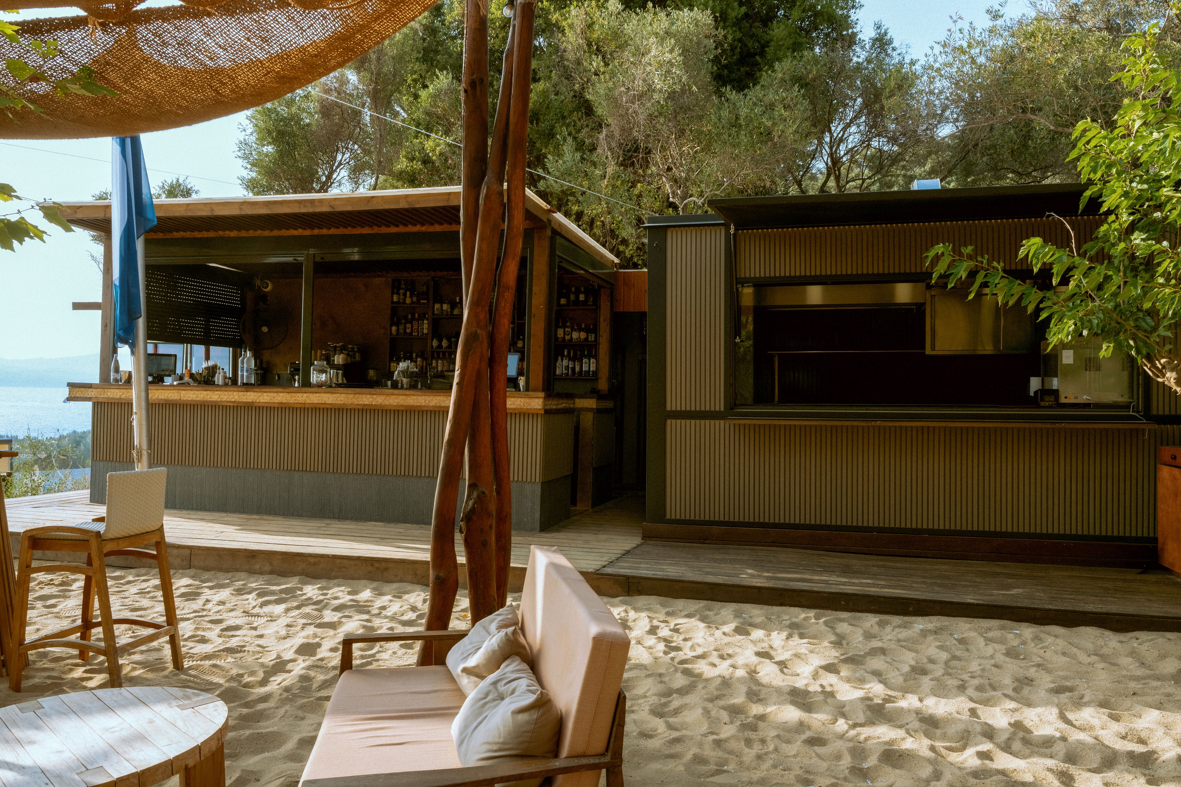 An inviting beach bar stands on sandy ground with wooden structures and a shaded canopy overhead. Comfortable cushioned seating is arranged in the foreground, facing the bar area stocked with bottles and glassware. Trees and greenery surround the open-air bar, creating a relaxed coastal atmosphere. Warm natural light enhances the casual, vacation-like setting.