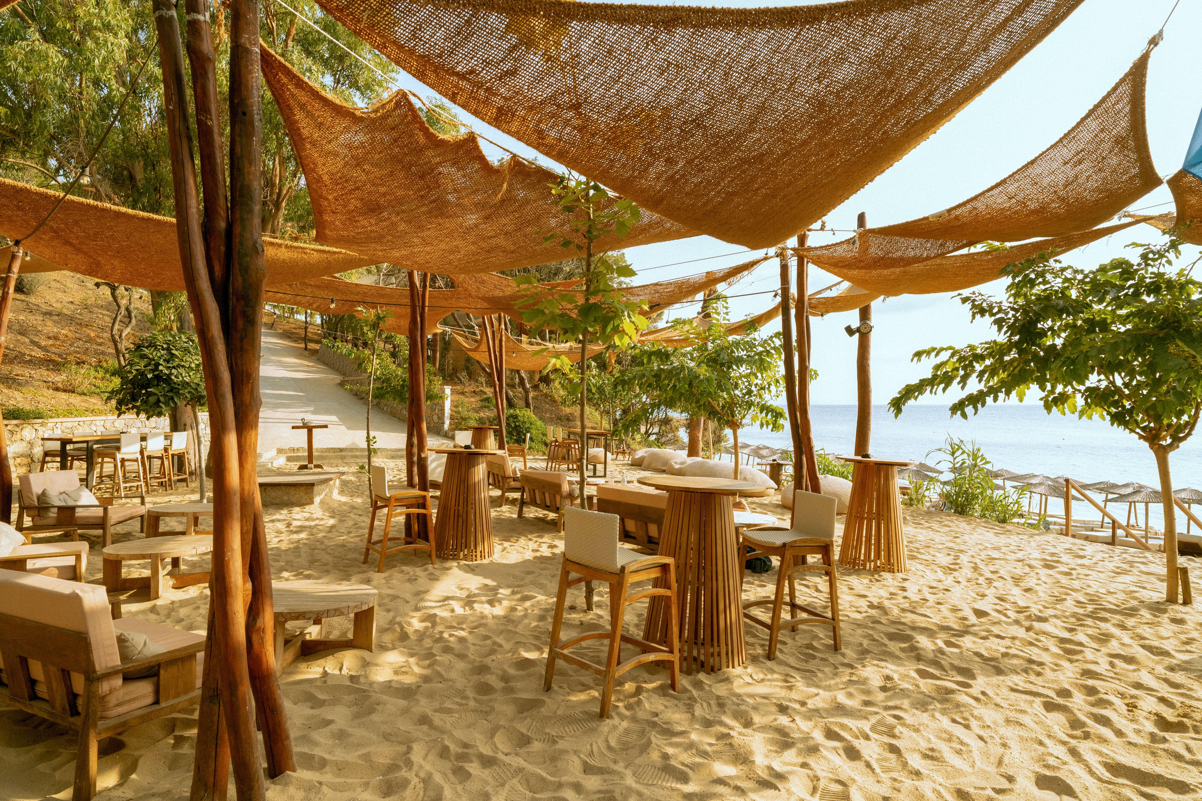 An open-air café is set directly on a sandy beach with wooden tables and cushioned chairs. Natural woven canopies are stretched overhead between wooden poles, creating shade and a warm, rustic atmosphere. Trees and greenery surround the seating area, and the shoreline with calm water is visible in the background. The scene suggests a relaxed coastal dining experience in a sunny, tropical setting.