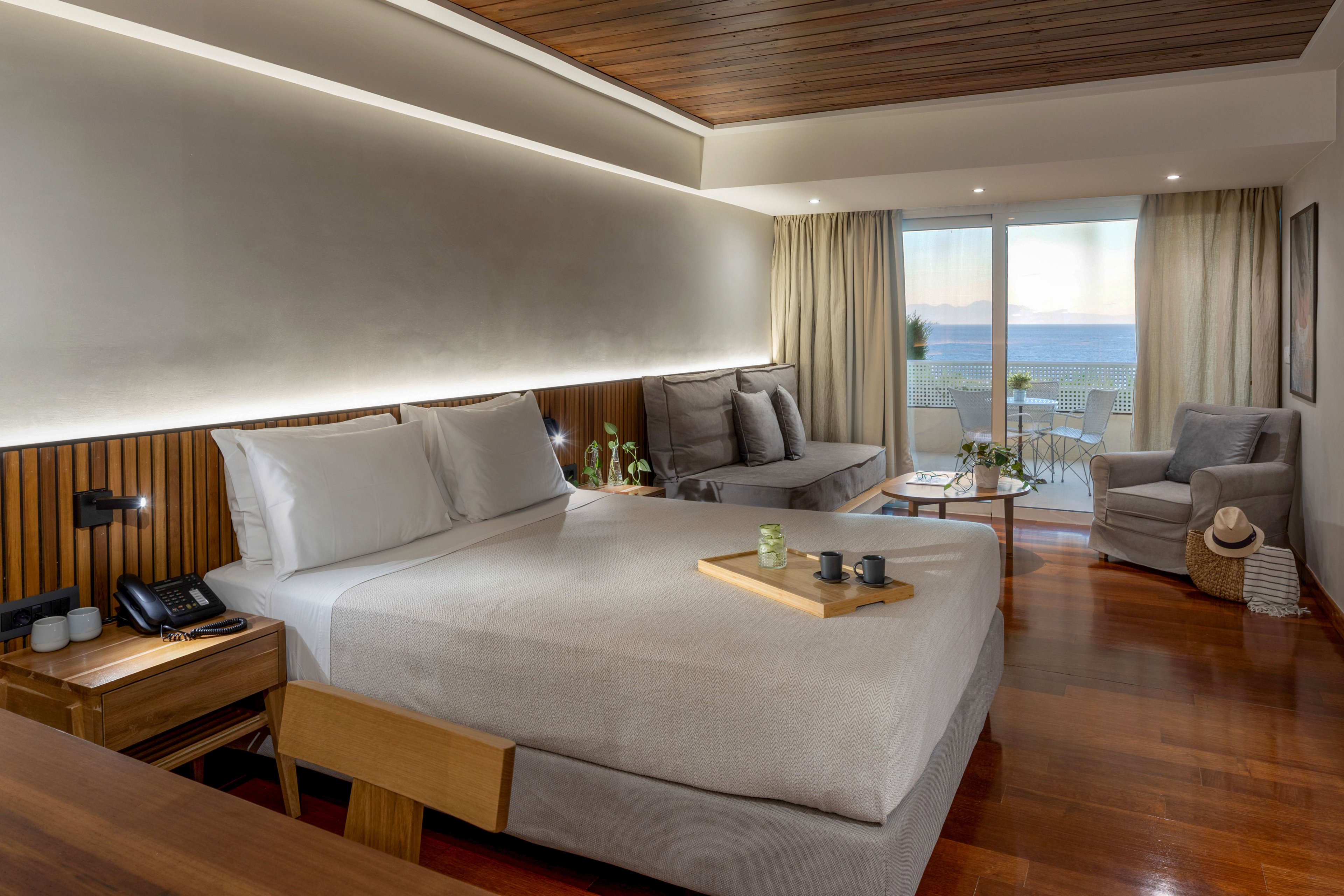 Contemporary hotel room features a large neatly made bed with neutral bedding and wooden furnishings. A tray with cups and a bottle sits on the bed, adding a touch of hospitality. The space includes a sofa, armchair, side tables, and soft ambient lighting along the headboard wall. Floor-to-ceiling glass doors open to a balcony with seating and a scenic ocean view in the distance.