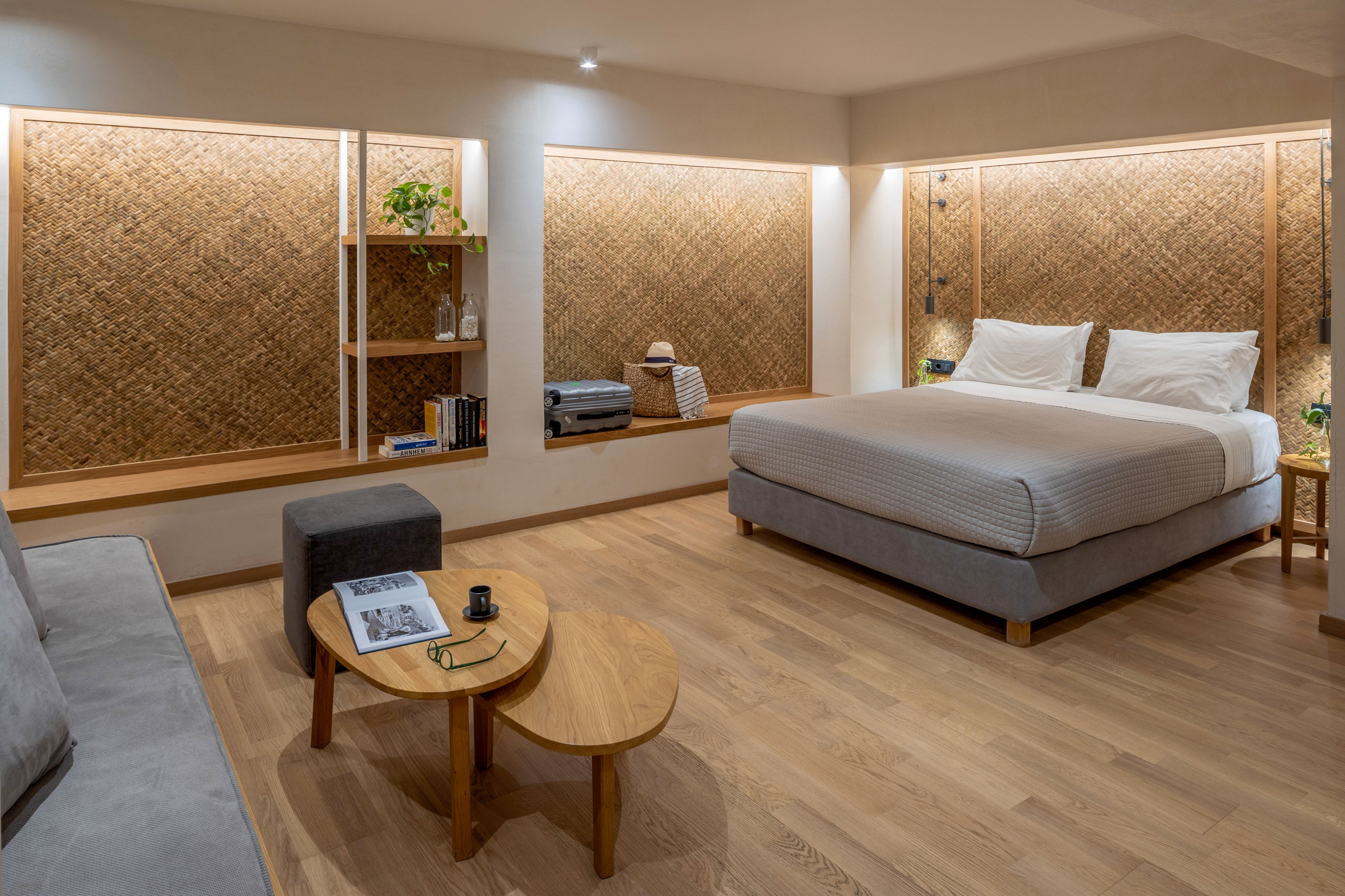 A modern minimalist bedroom features a large bed with white linens and neutral-toned upholstery. The room has warm wooden flooring and woven textured wall panels that frame built-in shelving. A small round wooden coffee table with a cup and saucer sits beside a gray ottoman and sofa. Soft ambient lighting creates a cozy, inviting atmosphere suitable for a hotel or contemporary home interior.