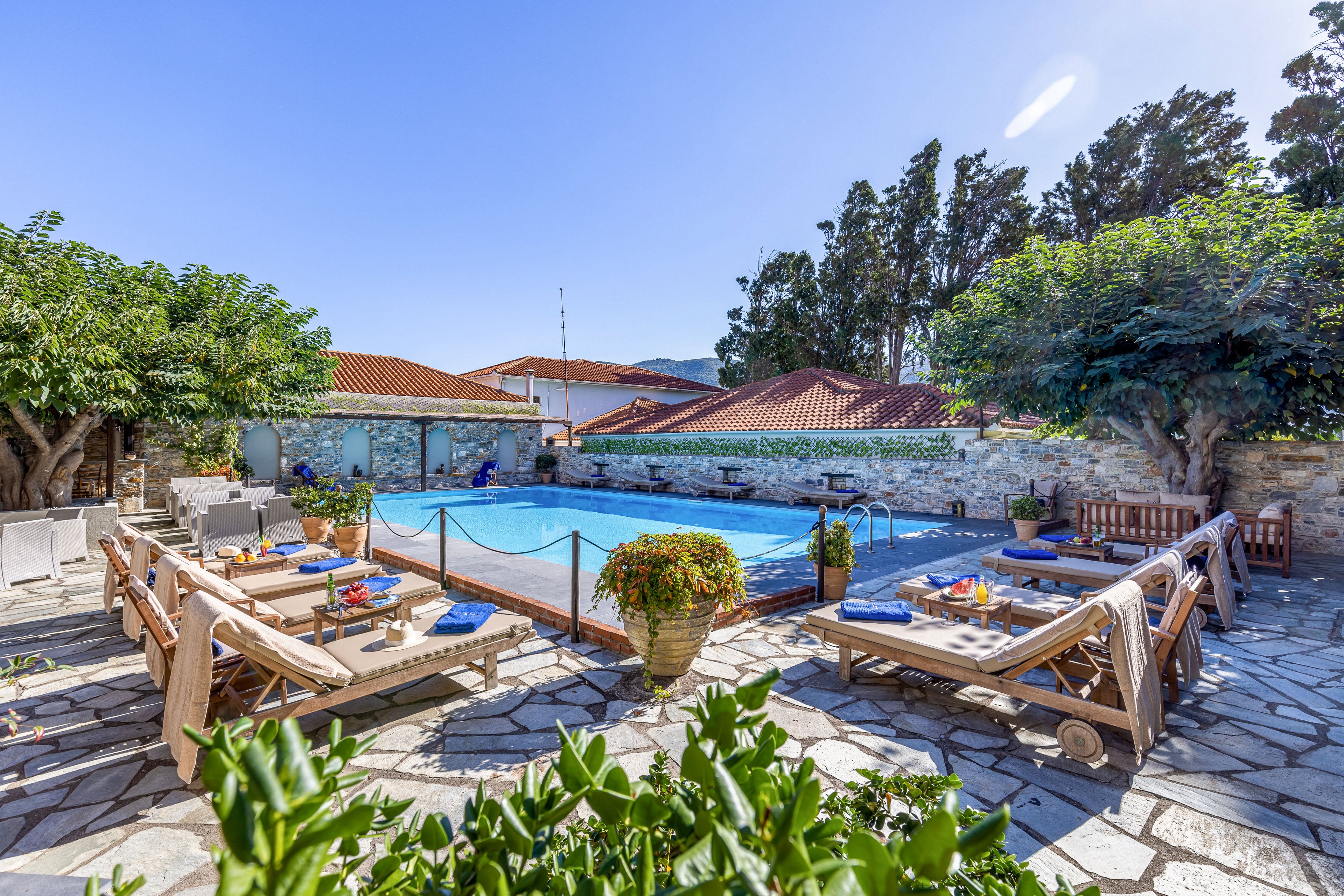 Outdoor swimming pool set in a stone courtyard with multiple sun loungers arranged around the water. Blue towels and cushions add color to the seating area, alongside small tables with drinks and decor. Lush green trees and potted plants frame the pool, while red-tiled roofs and a stone wall enclose the space. The bright daylight and clear blue sky create a relaxed vacation atmosphere.
