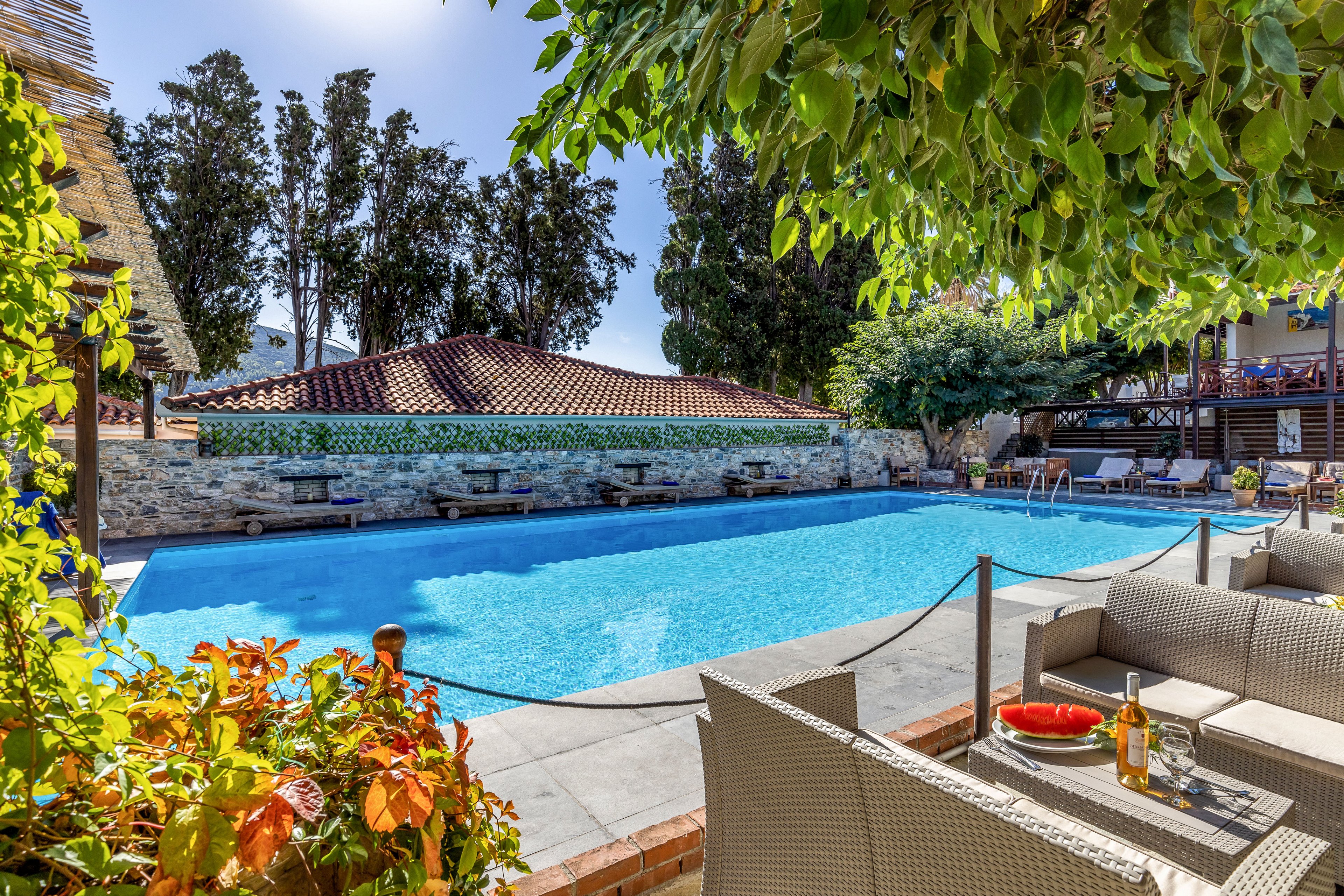 A bright outdoor swimming pool is surrounded by lounge chairs and shaded trees at a resort. In the foreground, a cozy wicker seating area holds a tray with a bottle of white wine and two glasses. The clear blue water reflects the sunny sky, creating a refreshing atmosphere. Stone walls, tiled roofs, and lush greenery frame the tranquil leisure setting.