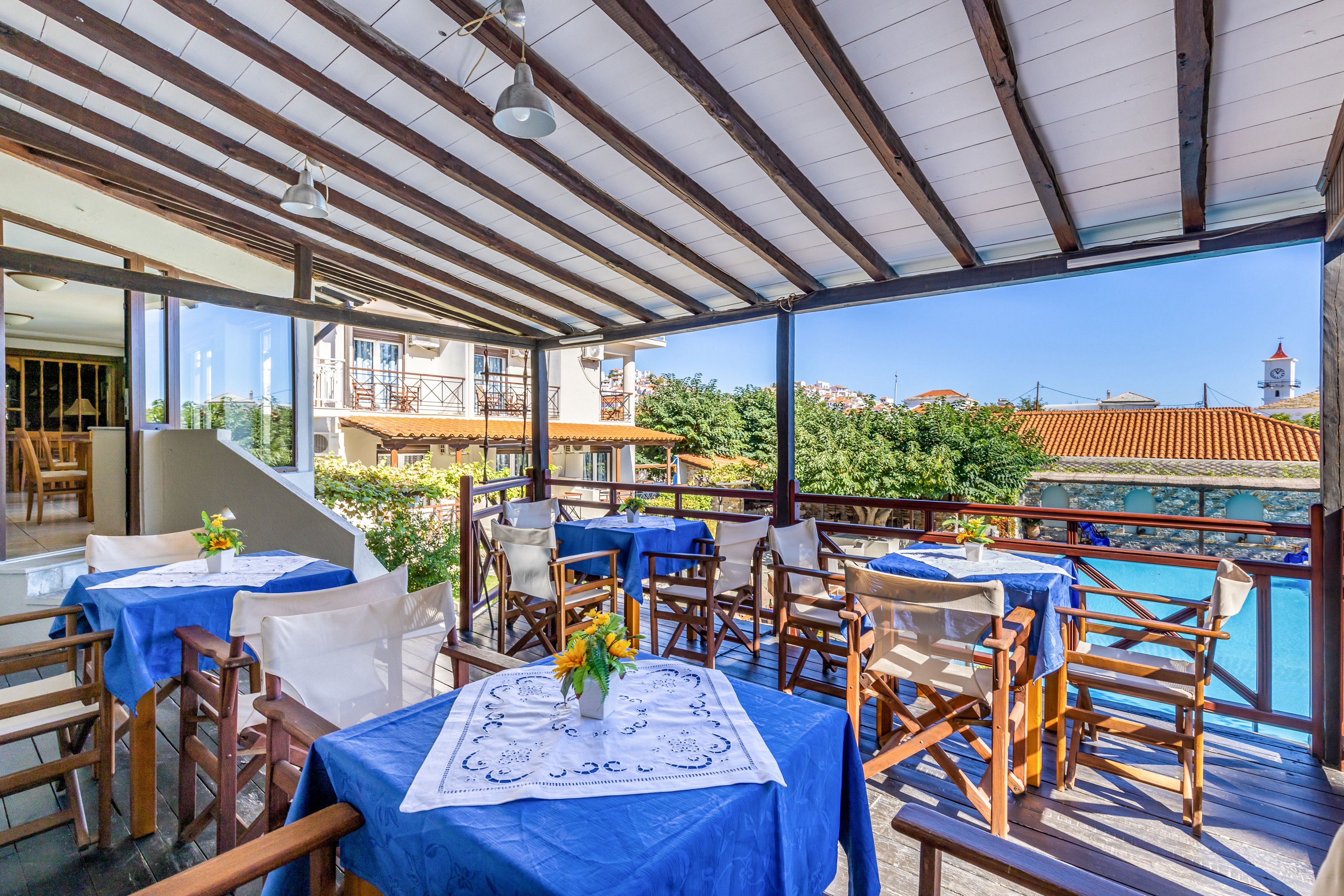 Open-air terrace restaurant overlooks a calm waterfront and village rooftops. Wooden tables are covered with bright blue cloths and white embroidered toppers, each decorated with a small vase of flowers. Wooden chairs with white cushions surround the tables under a slatted roof that filters the sunlight. In the background, trees, tiled roofs, and a distant tower complete the relaxed coastal setting.