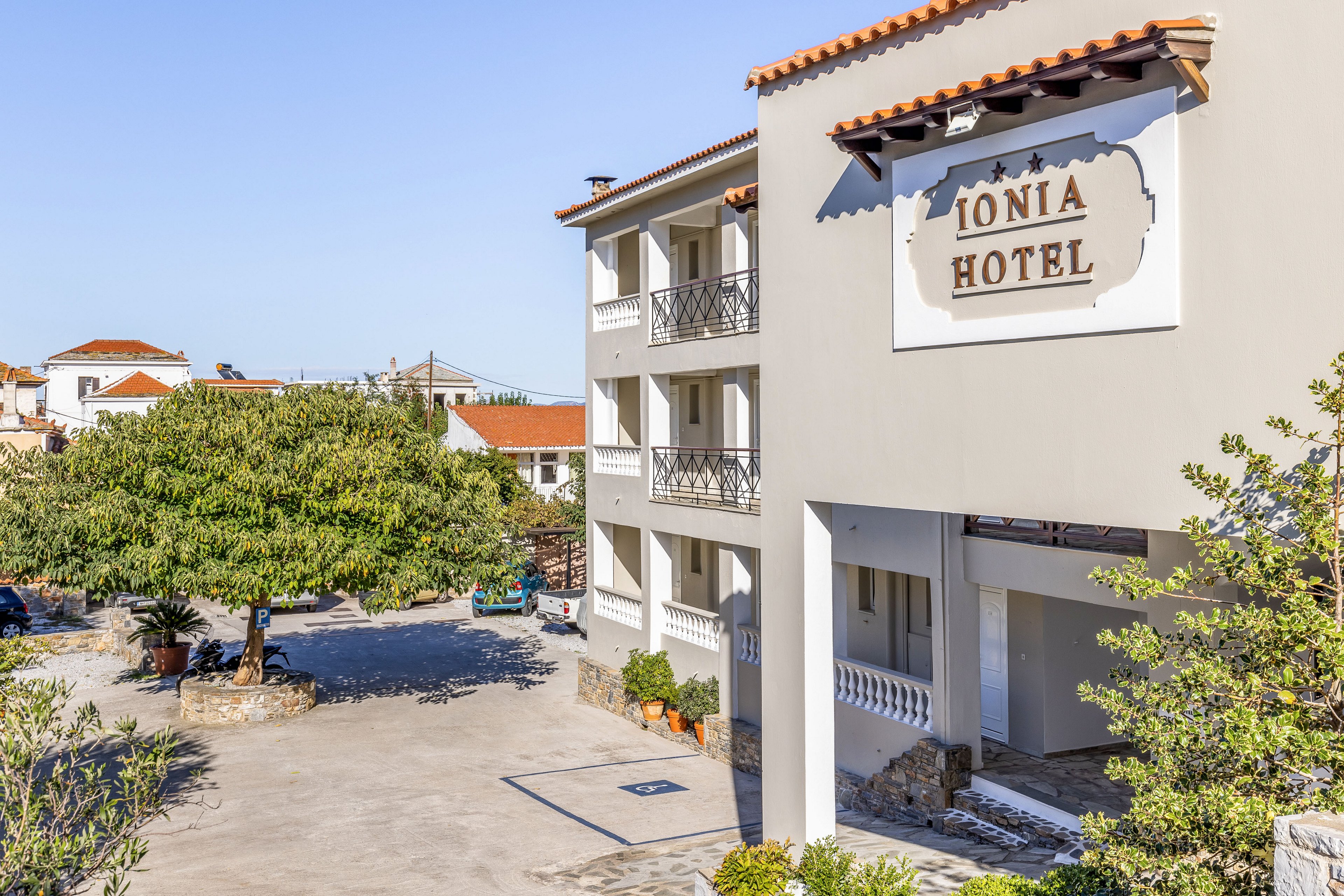 The image shows the exterior of the Ionia Hotel with a bright, sunlit courtyard. A multi-story building with balconies and tiled roof stands prominently on the right side. A large tree and surrounding greenery soften the paved open space, creating a welcoming atmosphere. Clear blue sky and distant rooftops suggest a warm, Mediterranean setting ideal for vacation stays.