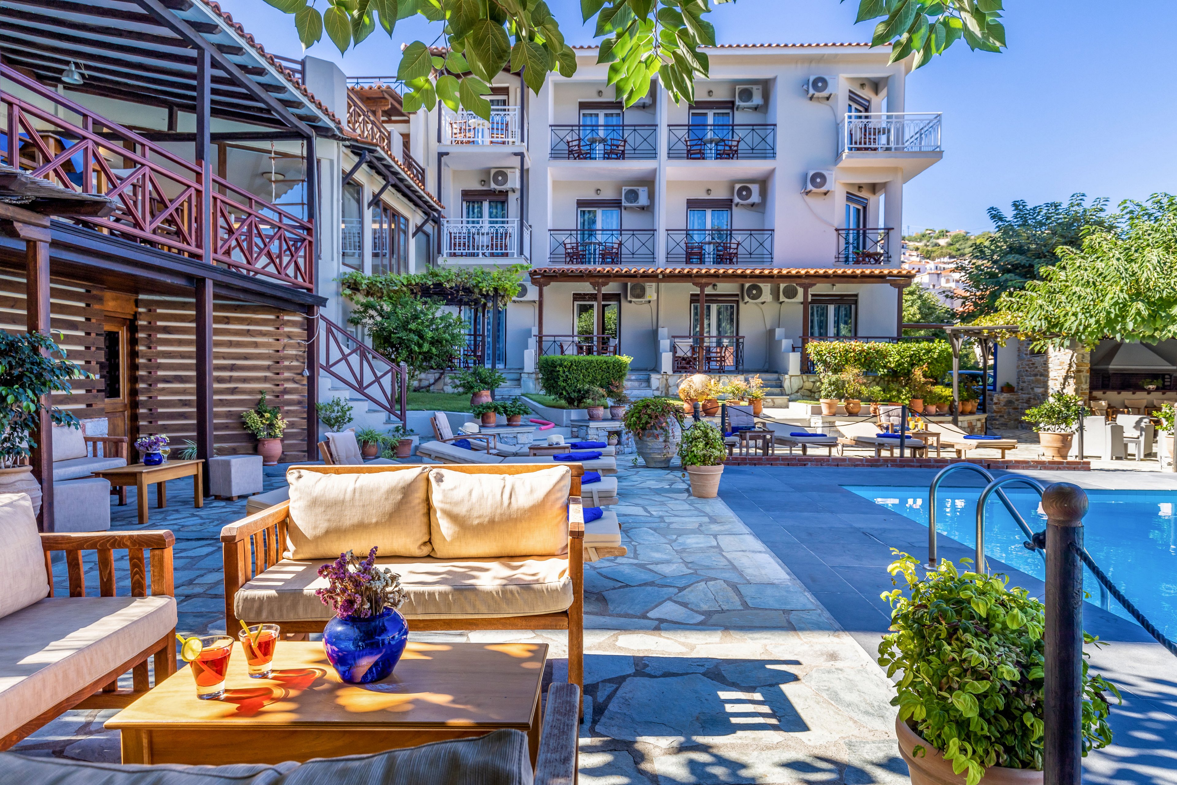 An inviting hotel courtyard features cushioned wooden seating around a bright blue swimming pool. A multi-story building with balconies and outdoor air-conditioning units rises in the background. Potted plants, trees, and colorful decor create a relaxed, vacation atmosphere. Sunlight highlights the stone patio and poolside lounge chairs, suggesting a warm, leisurely day.