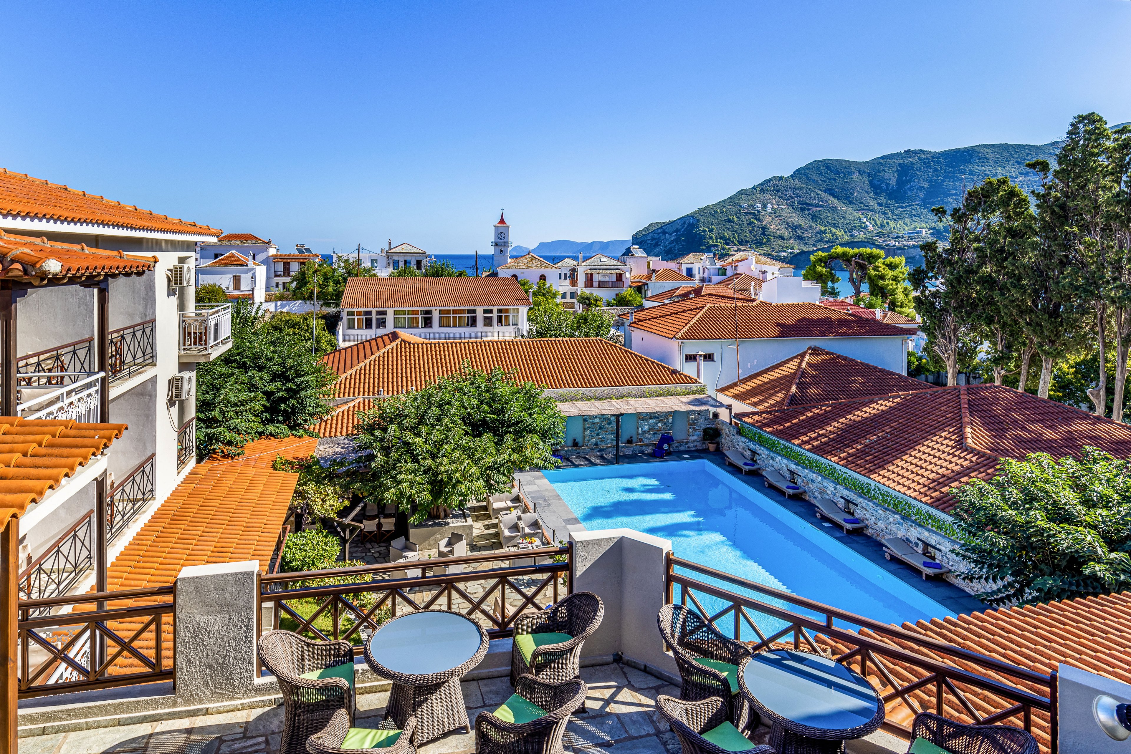 A sunny Mediterranean resort features a terrace with round tables and wicker chairs overlooking a central swimming pool. Traditional white buildings with red-tiled roofs surround the pool, framed by lush green trees. In the distance, the blue sea and a mountainous coastline create a scenic backdrop under a clear sky. The overall atmosphere is relaxed and inviting, ideal for vacation and leisure.