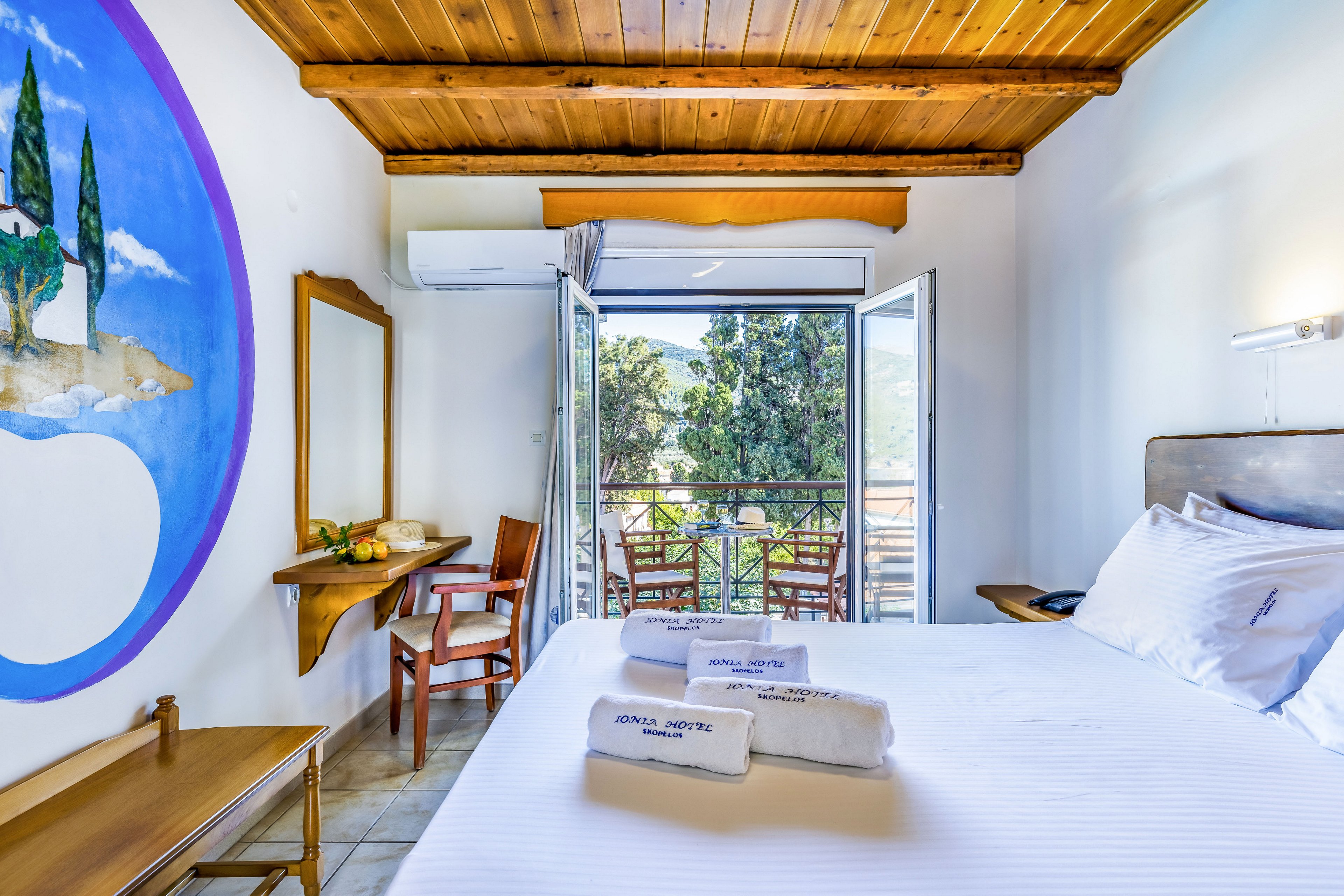 A neatly made hotel bed with folded white towels is positioned in a bright room featuring a wooden ceiling. The room opens onto a balcony with chairs and a view of lush trees and distant water. A colorful wall mural, wooden furniture, and a large mirror add warmth to the interior. Natural light floods the space through open glass doors, creating an inviting and relaxing atmosphere.