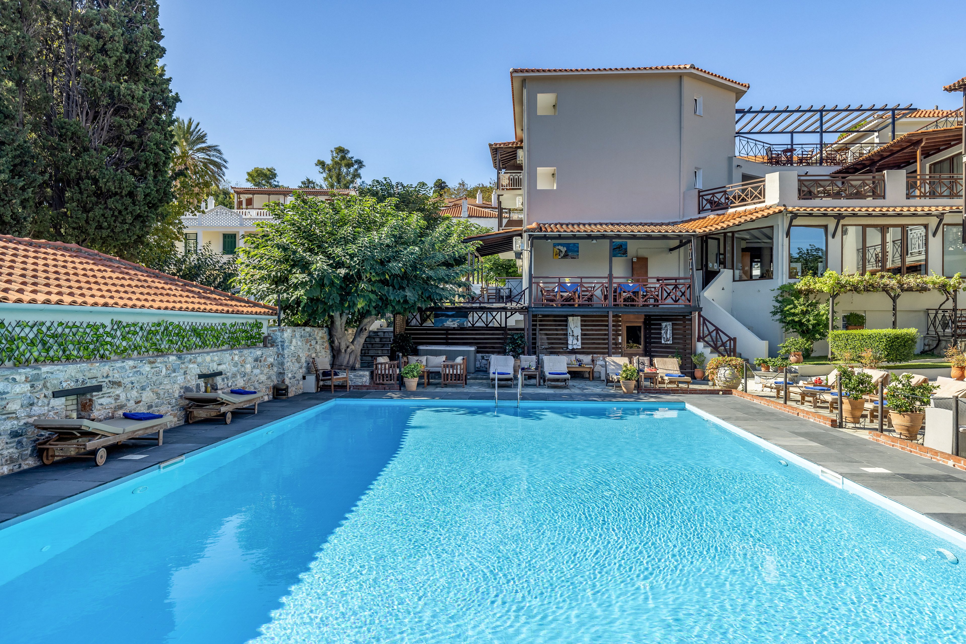 A clear blue swimming pool sits in front of a Mediterranean-style hotel complex. The buildings feature terracotta roof tiles, light-colored walls, and wooden balconies overlooking the water. Sun loungers and umbrellas line the poolside, creating a relaxed resort atmosphere. Lush greenery and trees surround the area, enhancing the tranquil vacation setting.