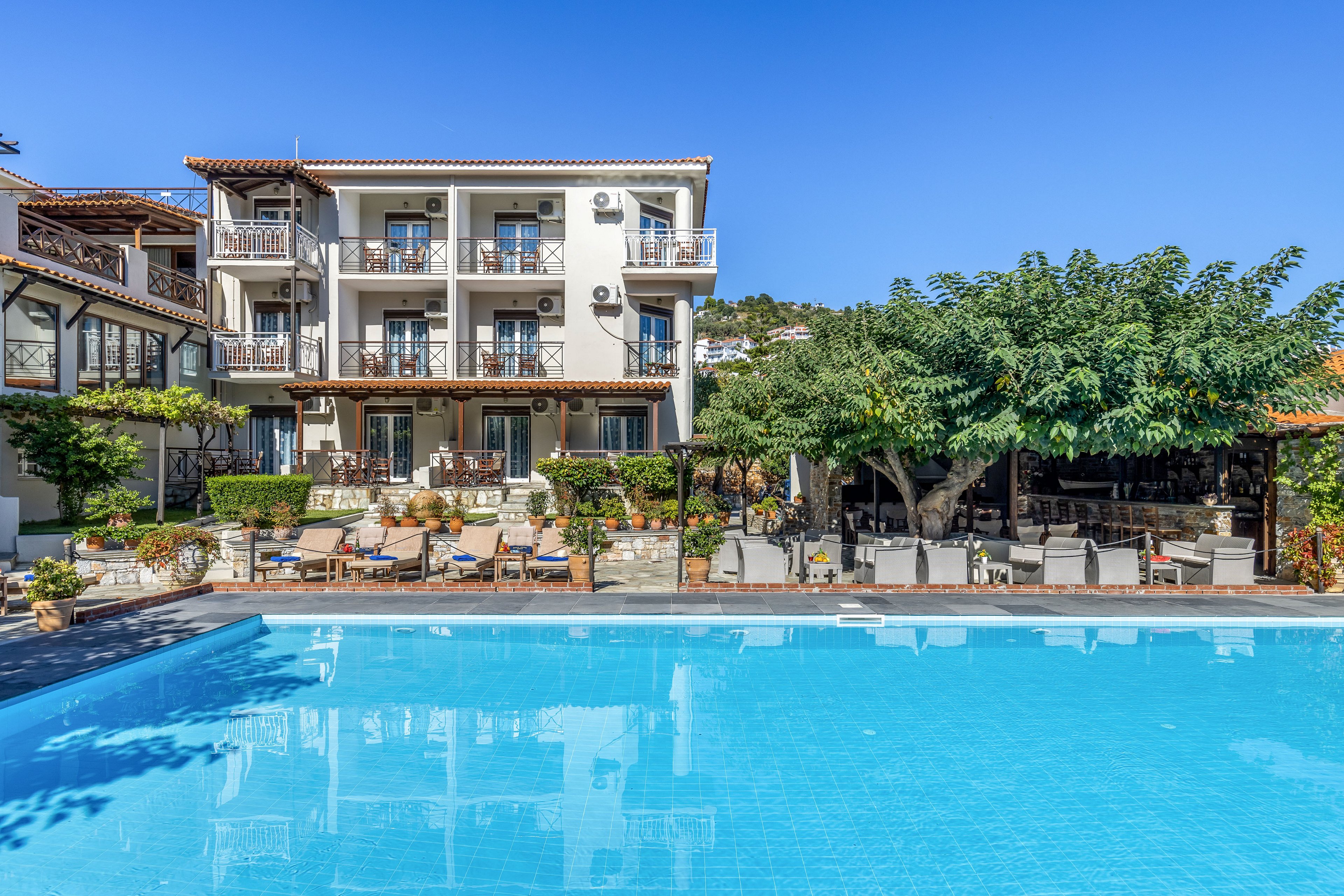A bright, multi-story hotel stands in front of a large outdoor swimming pool under a clear blue sky. The building features balconies with railings and outdoor seating areas overlooking the water. Sun loungers and tables are arranged around the poolside terrace, shaded by trees and pergolas. The scene suggests a relaxed vacation atmosphere in a warm, sunny destination.