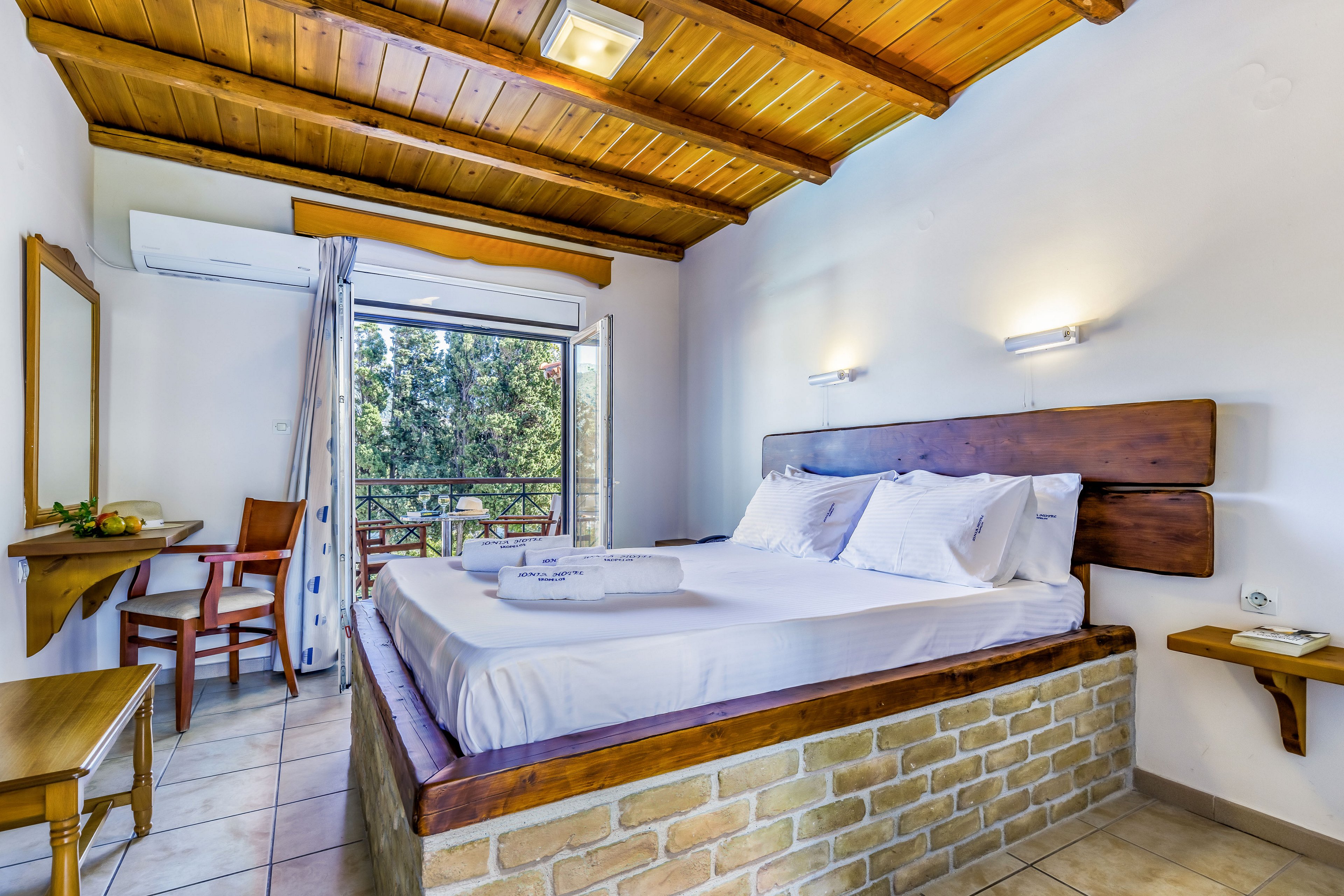 A cozy hotel bedroom features a large bed with white linens set on a brick and wood platform. The room has a wooden ceiling, tiled floor, and simple wooden furniture including a desk, chair, and wall mirror. An open balcony door reveals green trees outside, allowing natural light to fill the space. Wall-mounted lights and an air conditioning unit add comfort and functionality to the bright interior.