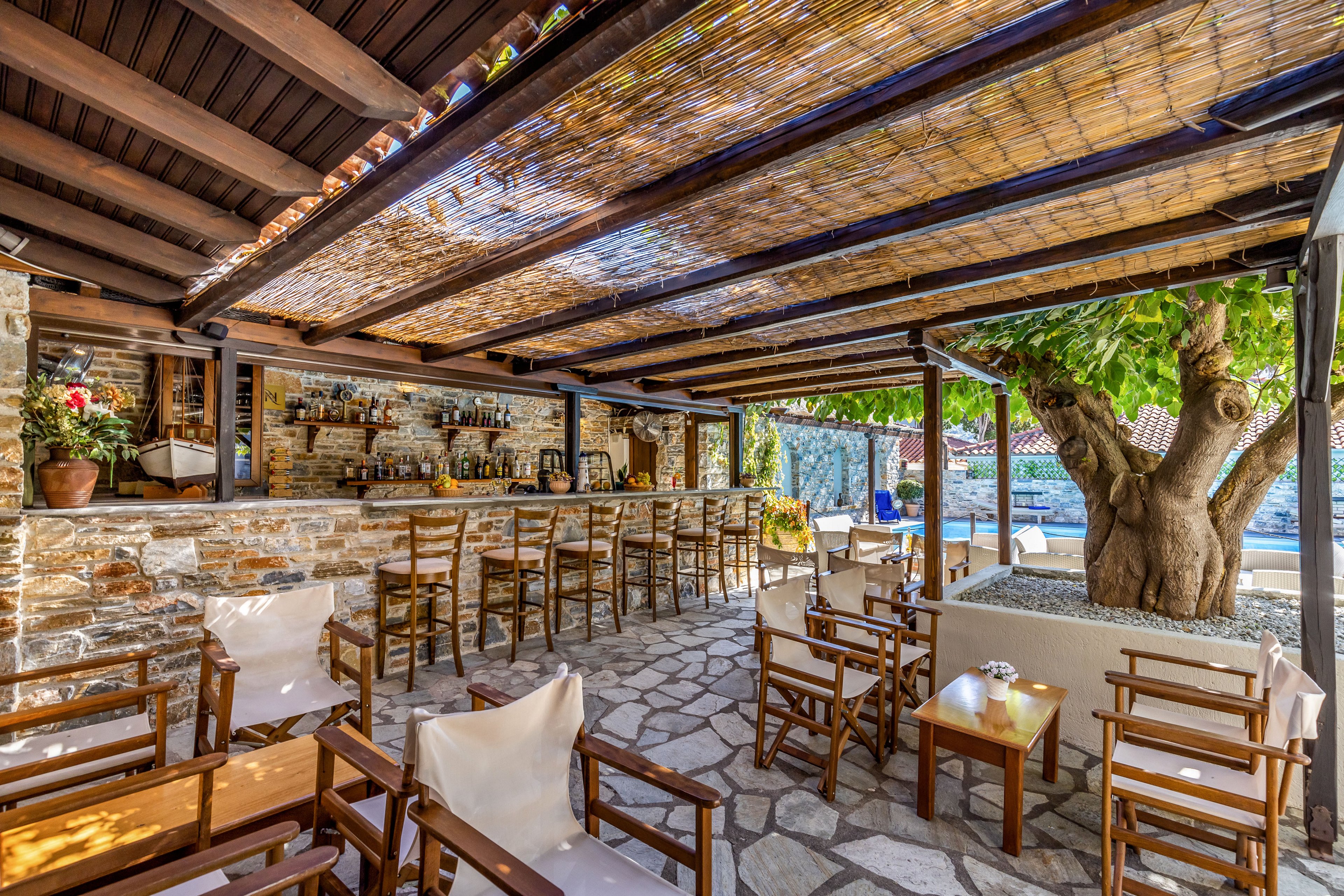 An open-air bar with a stone counter and wooden stools sits under a shaded pergola. Wooden chairs and small tables are arranged on a stone-paved floor, creating an inviting seating area. A large tree grows through a planter beside the bar, adding natural shade and character. Bottles and glassware line the back of the bar, suggesting a relaxed outdoor dining or cocktail setting in a warm climate.