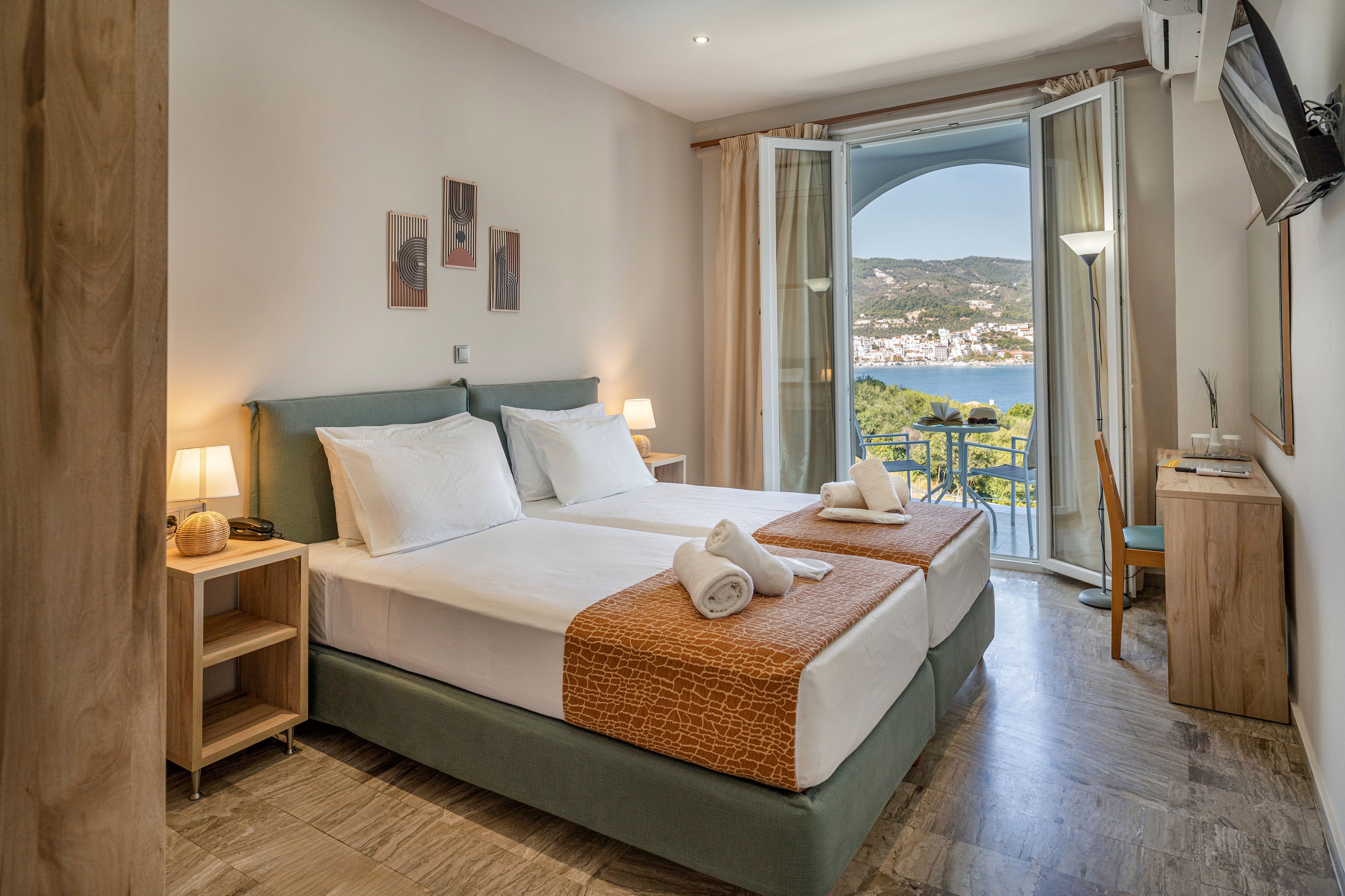 Bright, modern hotel room with two neatly made twin beds and folded towels on top. The space features warm wooden furniture, bedside lamps, and minimalist wall art. Large glass doors open to a balcony with chairs and a table overlooking a scenic bay and green hills. Natural light and neutral tones create a calm, inviting atmosphere ideal for vacation or relaxation.