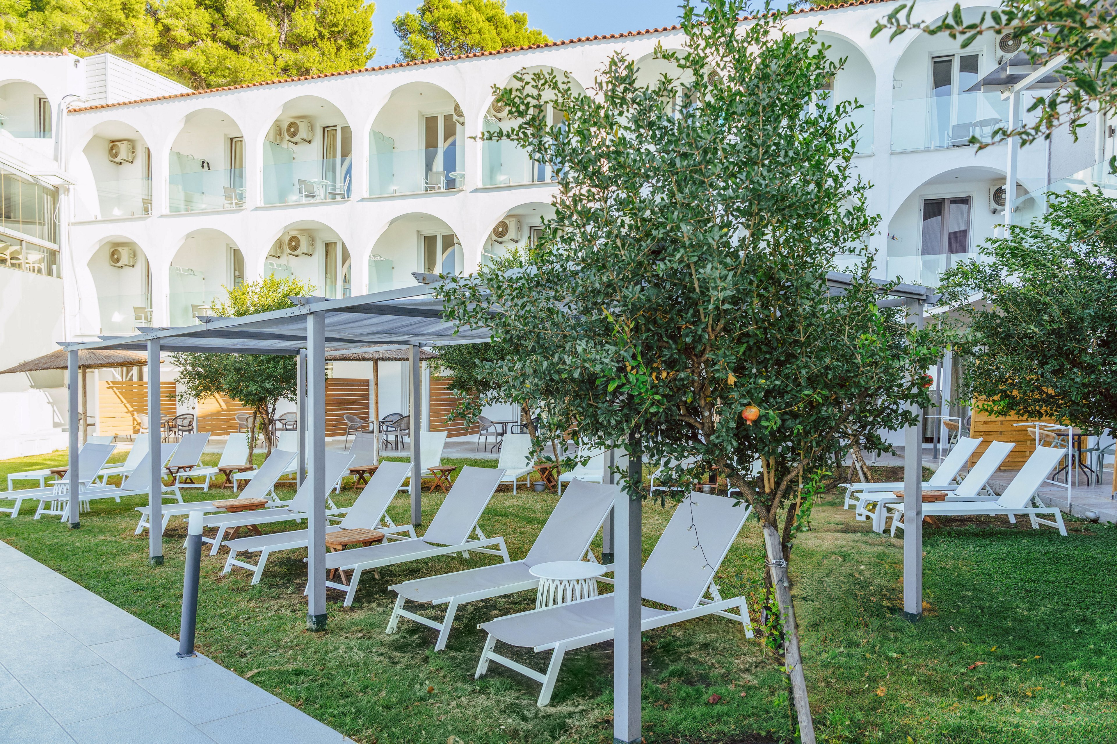 A modern white hotel building with arched balconies overlooks a neatly maintained garden. Numerous white sun loungers are arranged under simple pergola structures, creating a relaxing outdoor seating area. Green grass, small trees, and surrounding foliage add a fresh, natural atmosphere to the space. The scene suggests a peaceful vacation setting with comfortable outdoor amenities.