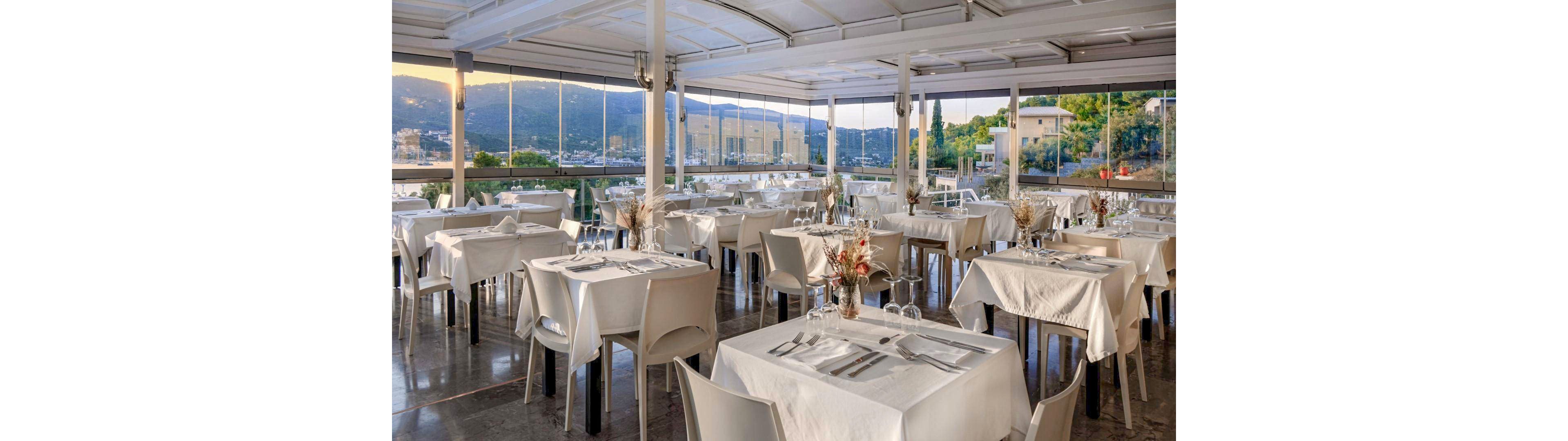 A bright, open-air restaurant terrace is set with neatly arranged white tablecloths and modern white chairs. Each table is prepared for dining with cutlery, glasses, and simple floral centerpieces. Large glass windows surround the space, revealing a scenic view of hills, buildings, and greenery in the distance. The polished floor and soft natural light create a clean, sophisticated atmosphere suitable for formal or casual dining.
