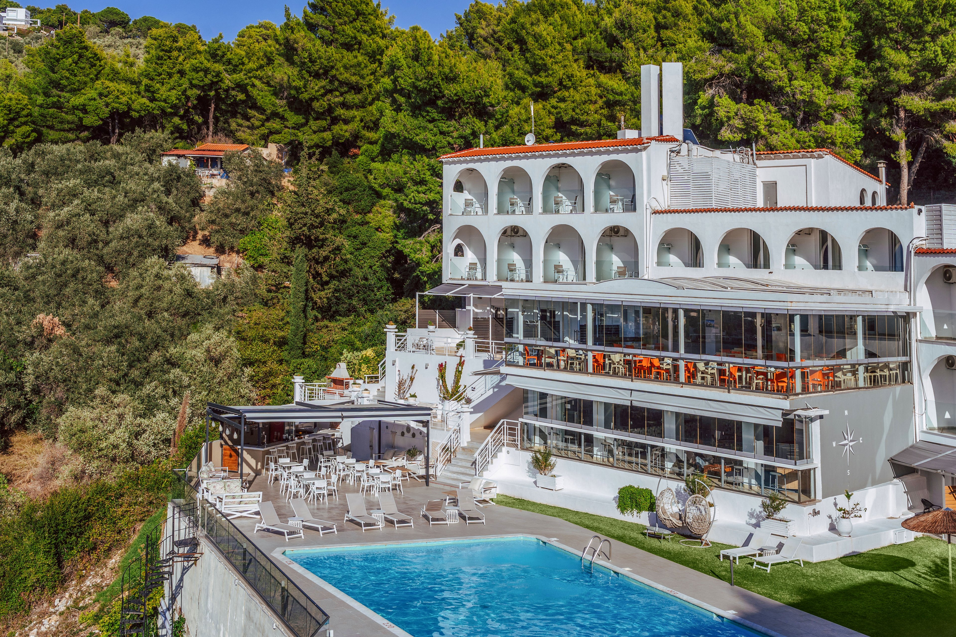 A white multi-story hotel stands on a hillside surrounded by lush green trees. The building features arched balconies and large glass windows overlooking an outdoor swimming pool. Sun loungers and tables are arranged around the pool and terrace areas, suggesting a relaxing vacation setting. The bright blue water and clear sky create a sunny, inviting atmosphere.