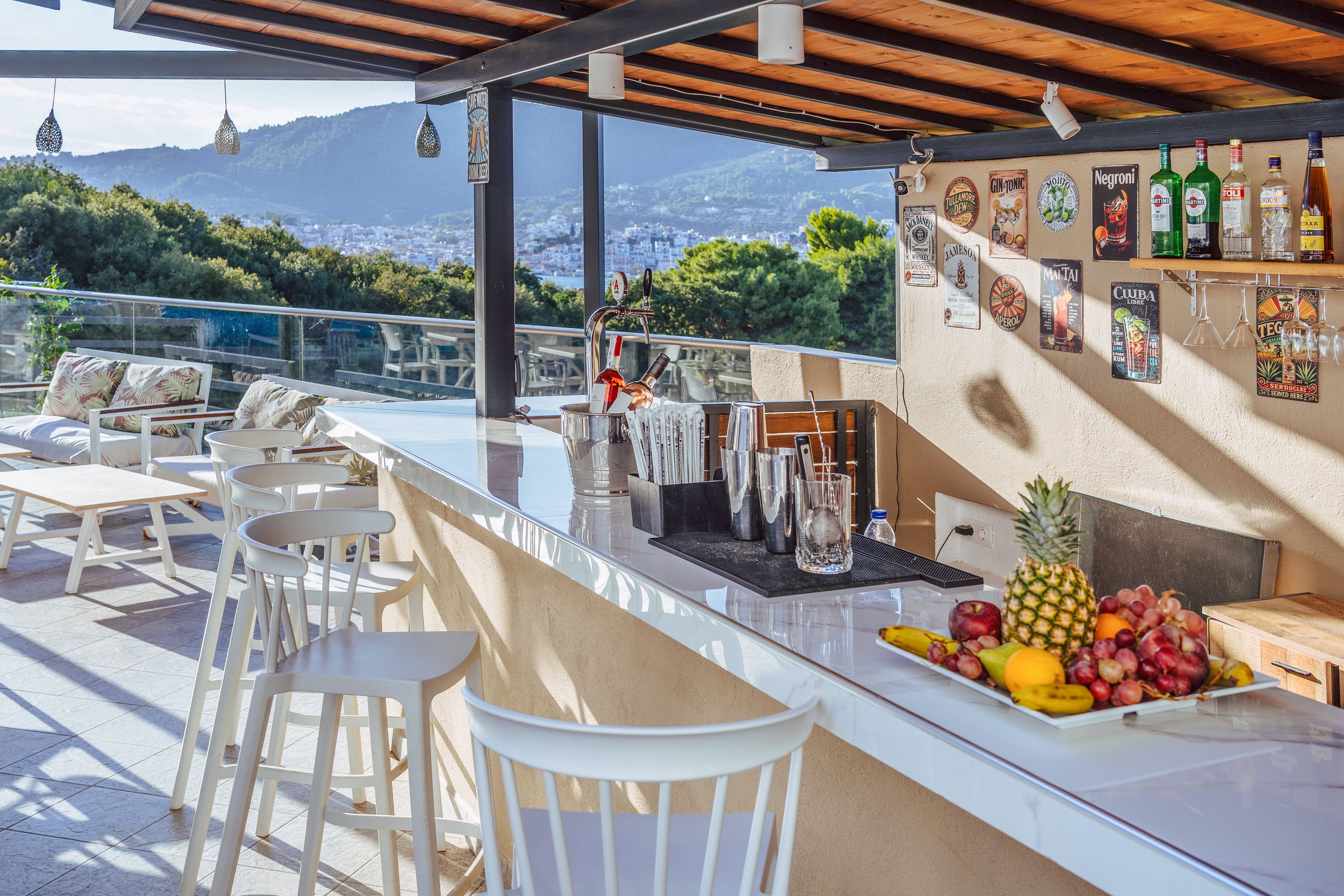 An open-air rooftop bar features a long white counter with stools and cocktail-making equipment. A colorful assortment of fresh fruits, including pineapple, bananas, and apples, is displayed on the bar. Shelves on the wall behind the counter hold various bottles and decorative drink posters. In the background, lounge seating and glass railings overlook a scenic hillside and cityscape under clear daylight.