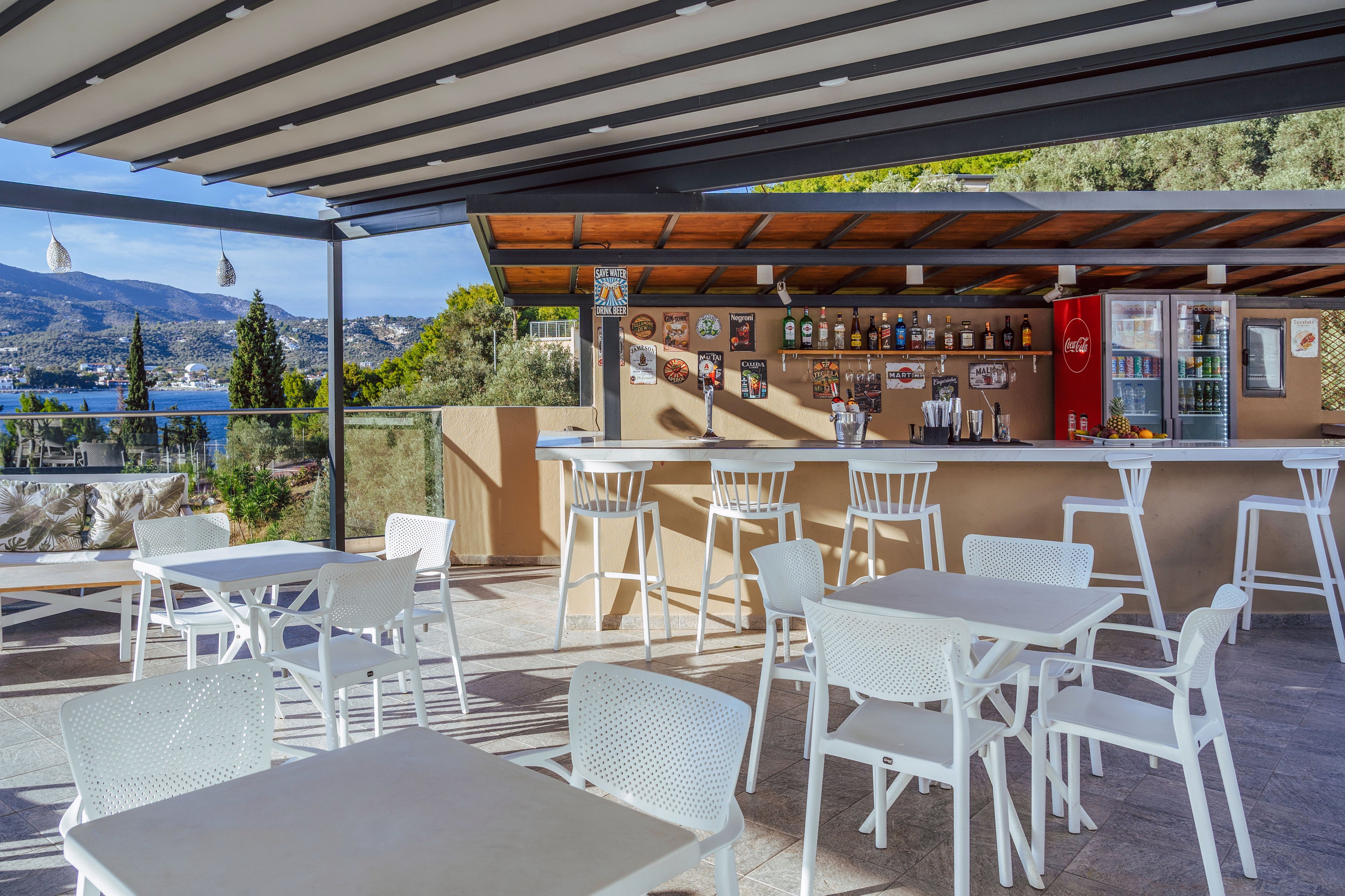An open-air bar terrace features white tables and chairs under a modern pergola roof. The bar counter in the background is stocked with bottles, glasses, and a red refrigerator, with decorative photos and signs on the wall. Large glass railings frame a scenic coastal landscape with blue water, hills, and greenery. Bright daylight and clean contemporary furniture create a relaxed holiday atmosphere.