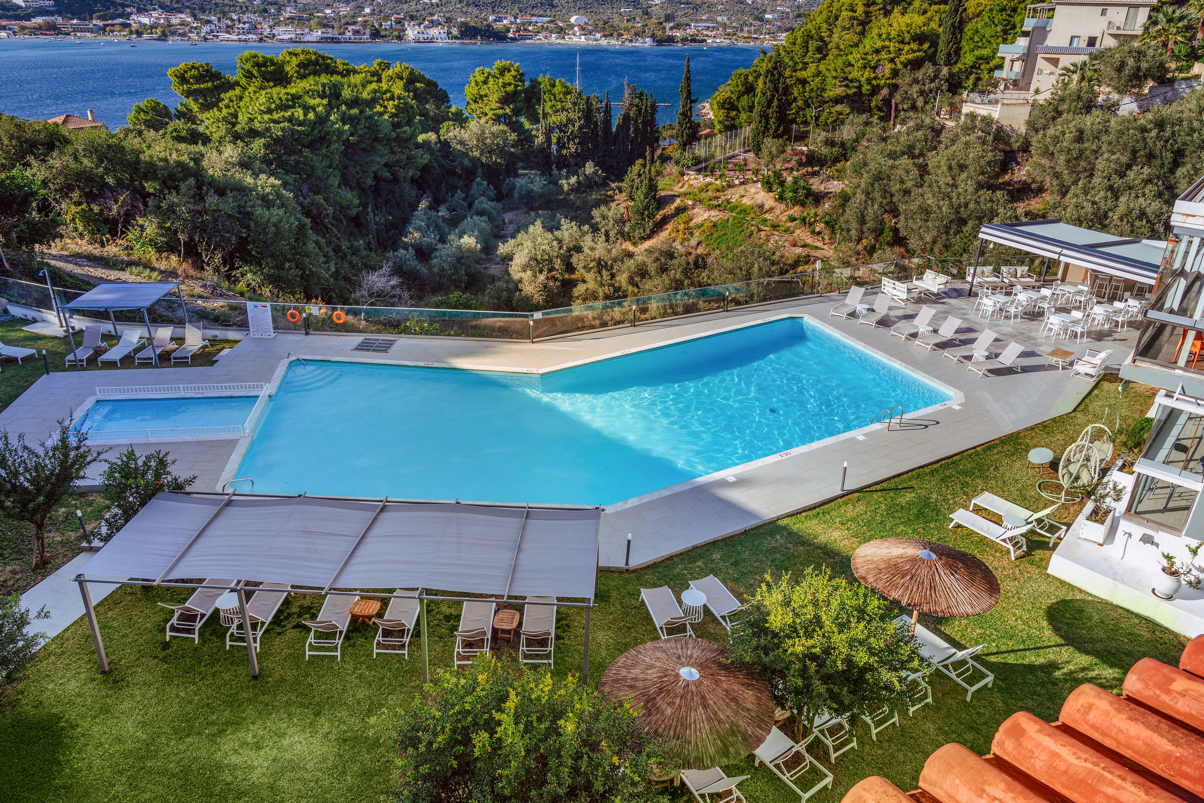 An elevated view shows a large outdoor swimming pool with an adjacent smaller pool surrounded by sun loungers and umbrellas. The pool area is part of a modern hotel complex set on a hillside above a scenic coastal bay. Green lawns, shaded pergolas, and seating areas create a relaxing resort atmosphere. In the background, dense trees and distant buildings line the shoreline beside the calm blue water.