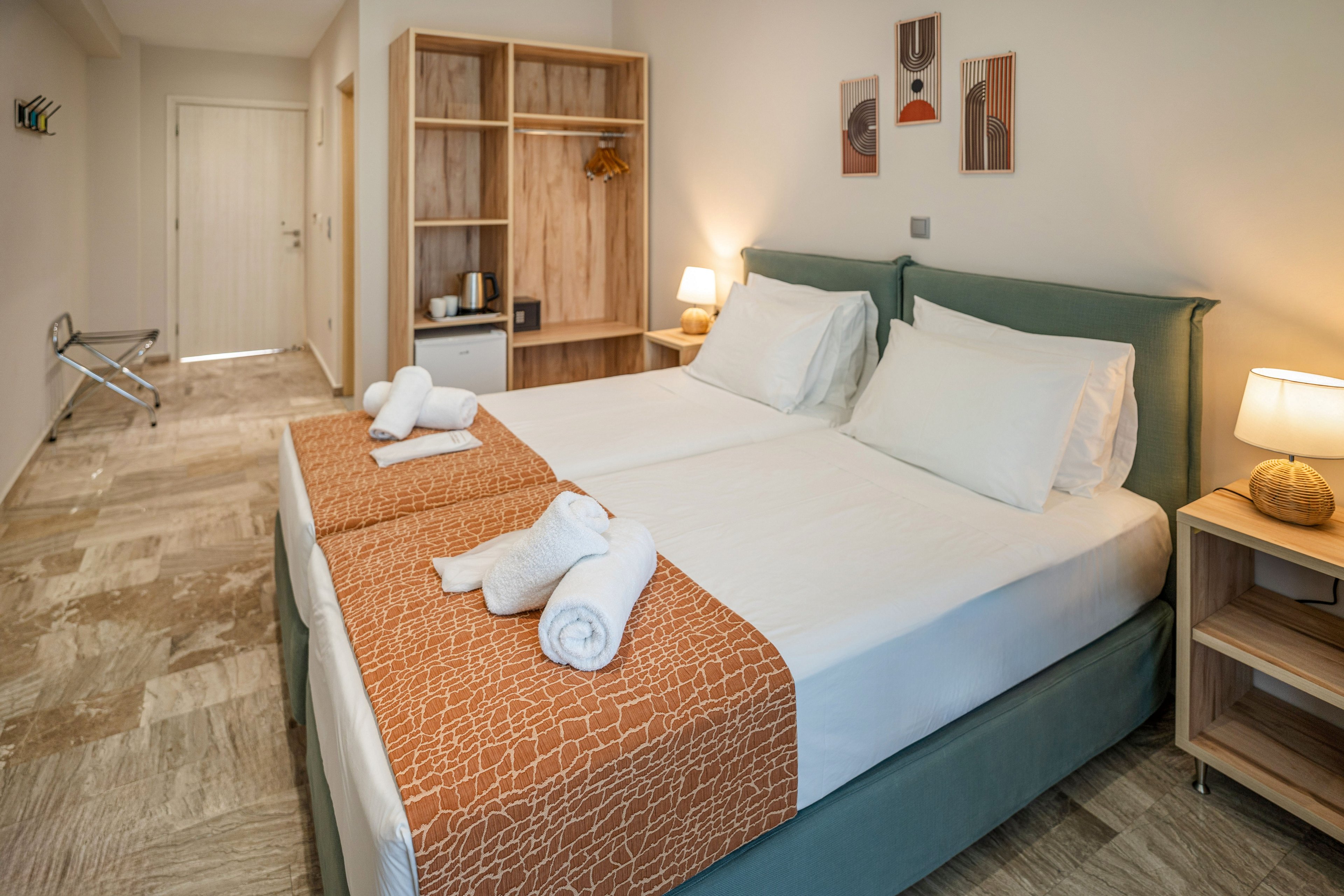 A contemporary hotel room features two neatly made twin beds with white linens and patterned orange runners. Rolled white towels are placed on each bed, adding a welcoming touch. The room includes a wooden open wardrobe with a kettle and mini fridge, a bedside table with a lit lamp, and abstract wall art above the headboard. Neutral tones and clean lines create a calm, minimalist atmosphere in the bright interior.