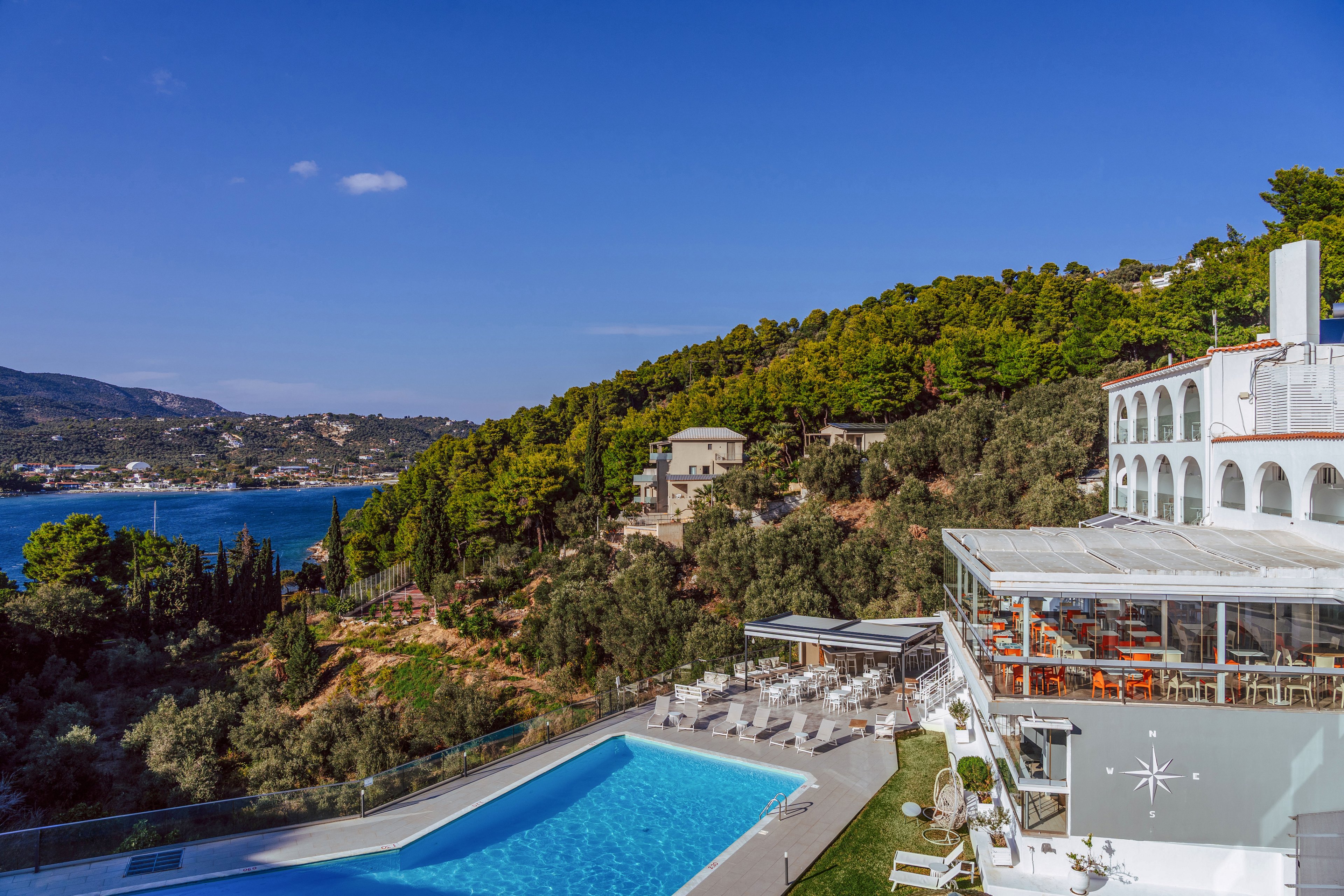 A modern seaside resort sits on a hillside overlooking a calm blue bay. In the foreground, a large outdoor swimming pool is surrounded by sun loungers and terraces. The white building features arched windows and a glass-enclosed dining area with orange chairs. Lush green trees cover the surrounding slopes, leading down to the water and distant houses along the coastline. The clear sky and bright sunlight create a vibrant, relaxing vacation atmosphere.