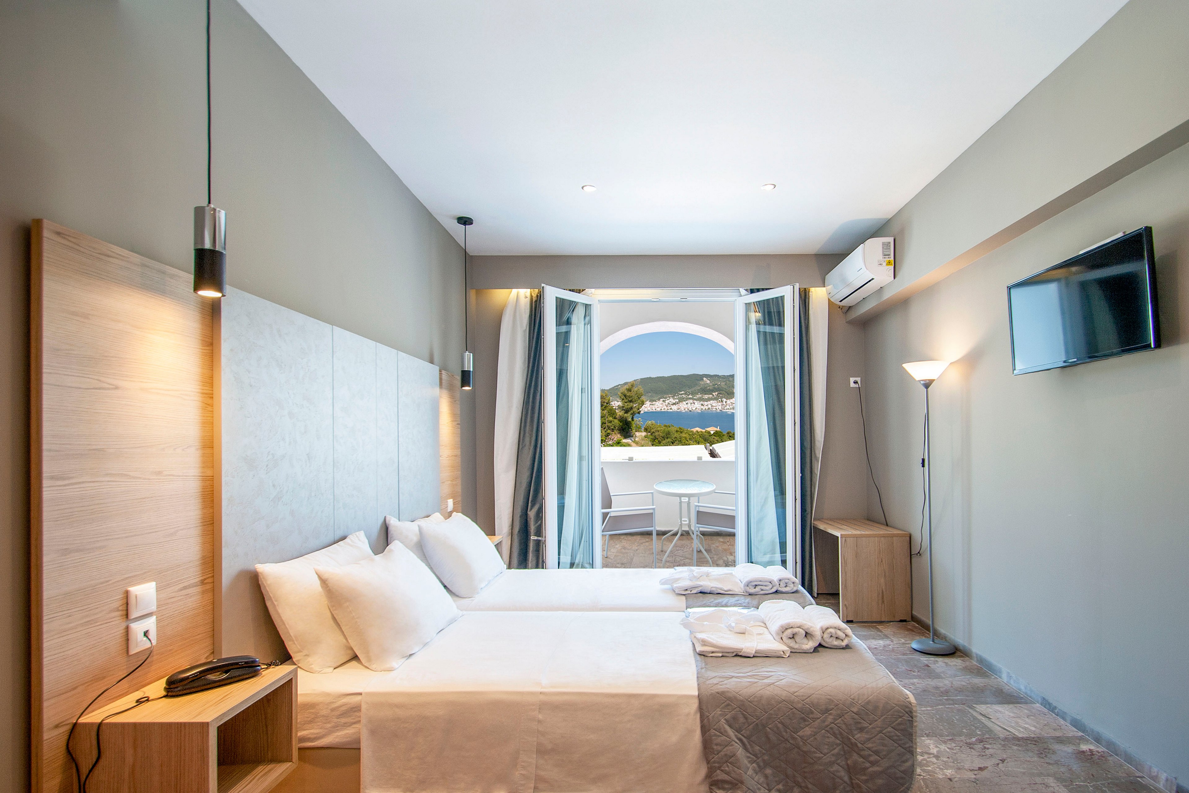 A contemporary hotel room features two neatly made single beds with white linens and pillows. Soft neutral walls and light wood furniture create a clean, minimalist aesthetic, complemented by pendant and floor lamps. Open glass doors lead to a balcony with a table and chairs, overlooking a scenic coastal landscape and hills. A wall-mounted flat-screen television and air conditioning unit complete the comfortable, vacation-ready space.