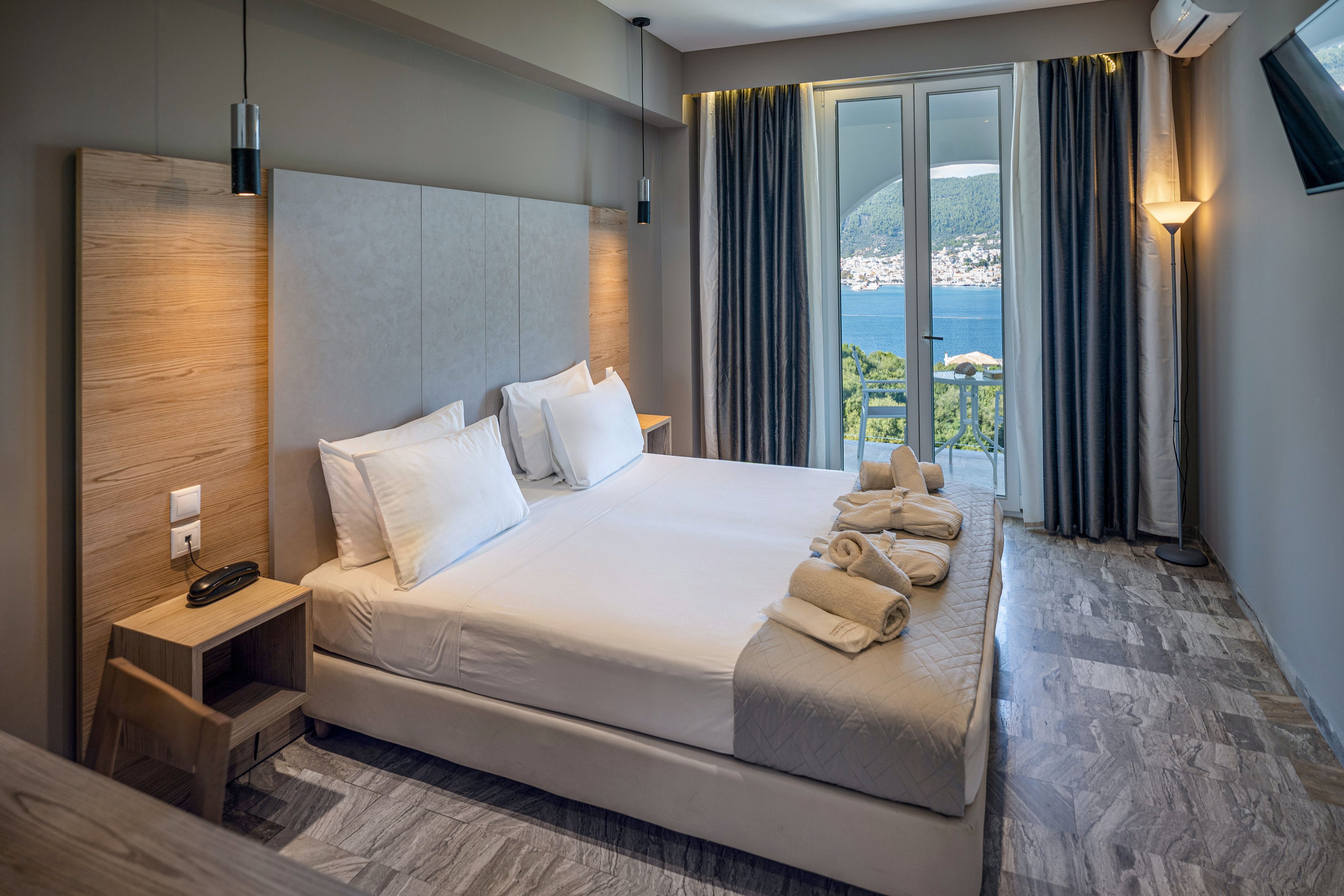 A contemporary hotel bedroom features a large double bed neatly made with white linens and pillows. Rolled towels are arranged on the bed, suggesting readiness for guests. Floor-to-ceiling glass doors open to a balcony overlooking a scenic coastal landscape with hills and water. Neutral tones, soft lighting, and minimalist furniture create a calm, relaxing atmosphere.