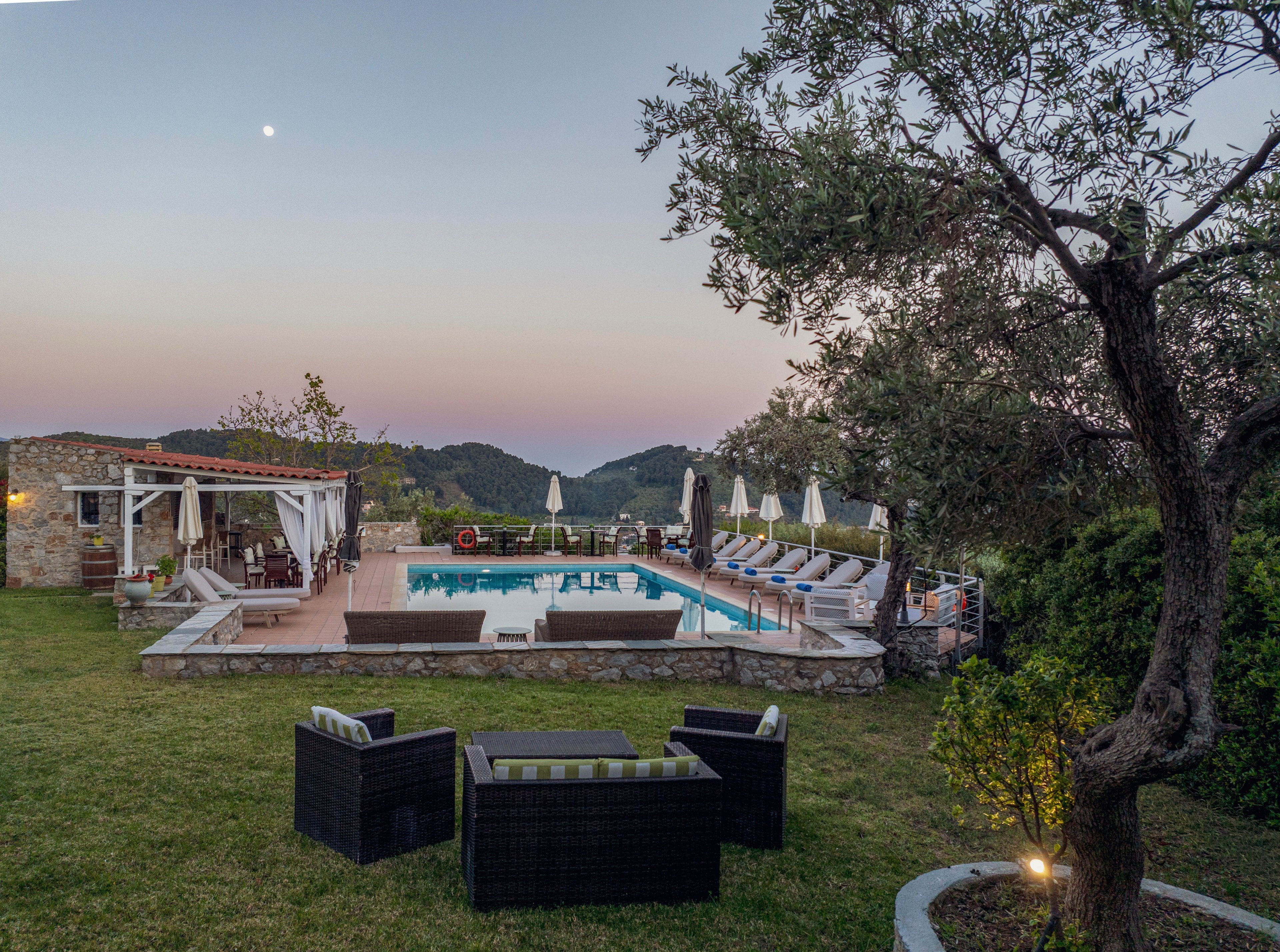 Outdoor swimming pool area at a hilltop villa during sunset. Lounge chairs and white umbrellas line the poolside terrace, with a small open-air bar or dining area to the left. In the foreground, dark wicker patio furniture sits on a manicured lawn beneath mature trees. Distant hills and a pastel evening sky with a visible moon create a tranquil vacation atmosphere.