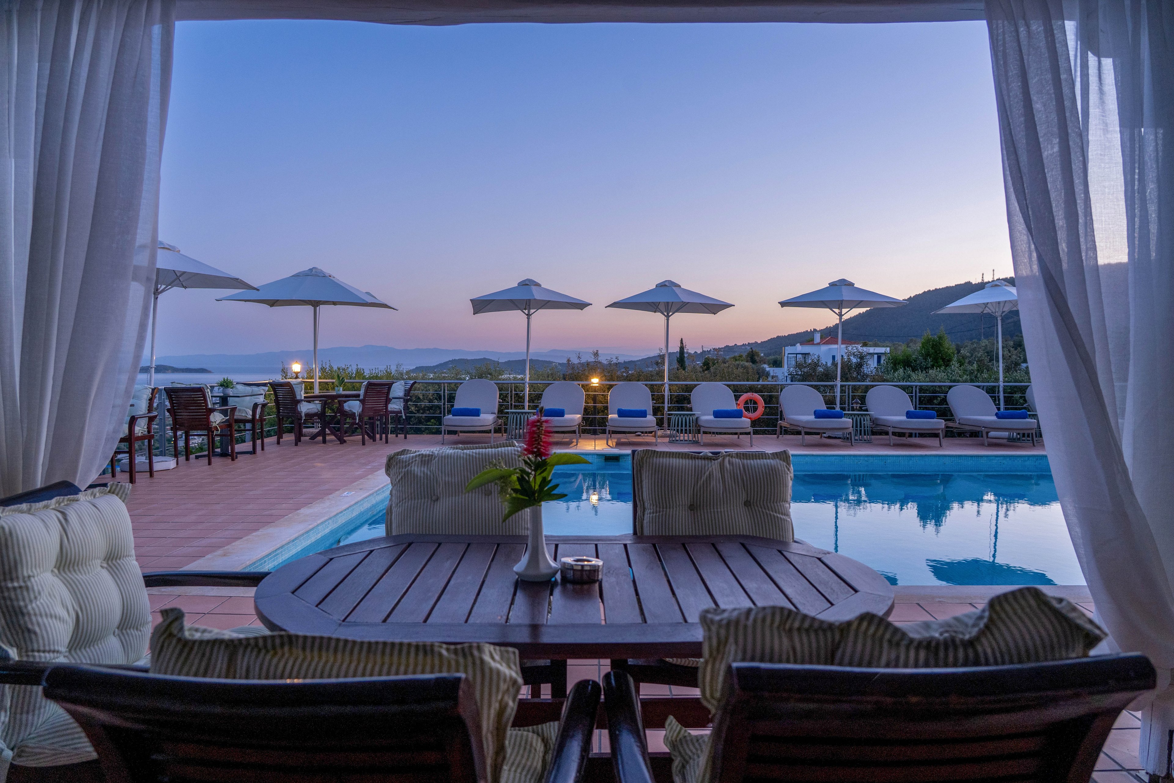 An elegant outdoor lounge area overlooks a tranquil swimming pool at dusk. Cushioned chairs surround a round wooden table framed by sheer white curtains. Sun loungers and white umbrellas line the pool deck, reflecting softly in the calm water. The sky glows with pastel evening colors over distant hills, creating a serene vacation atmosphere.