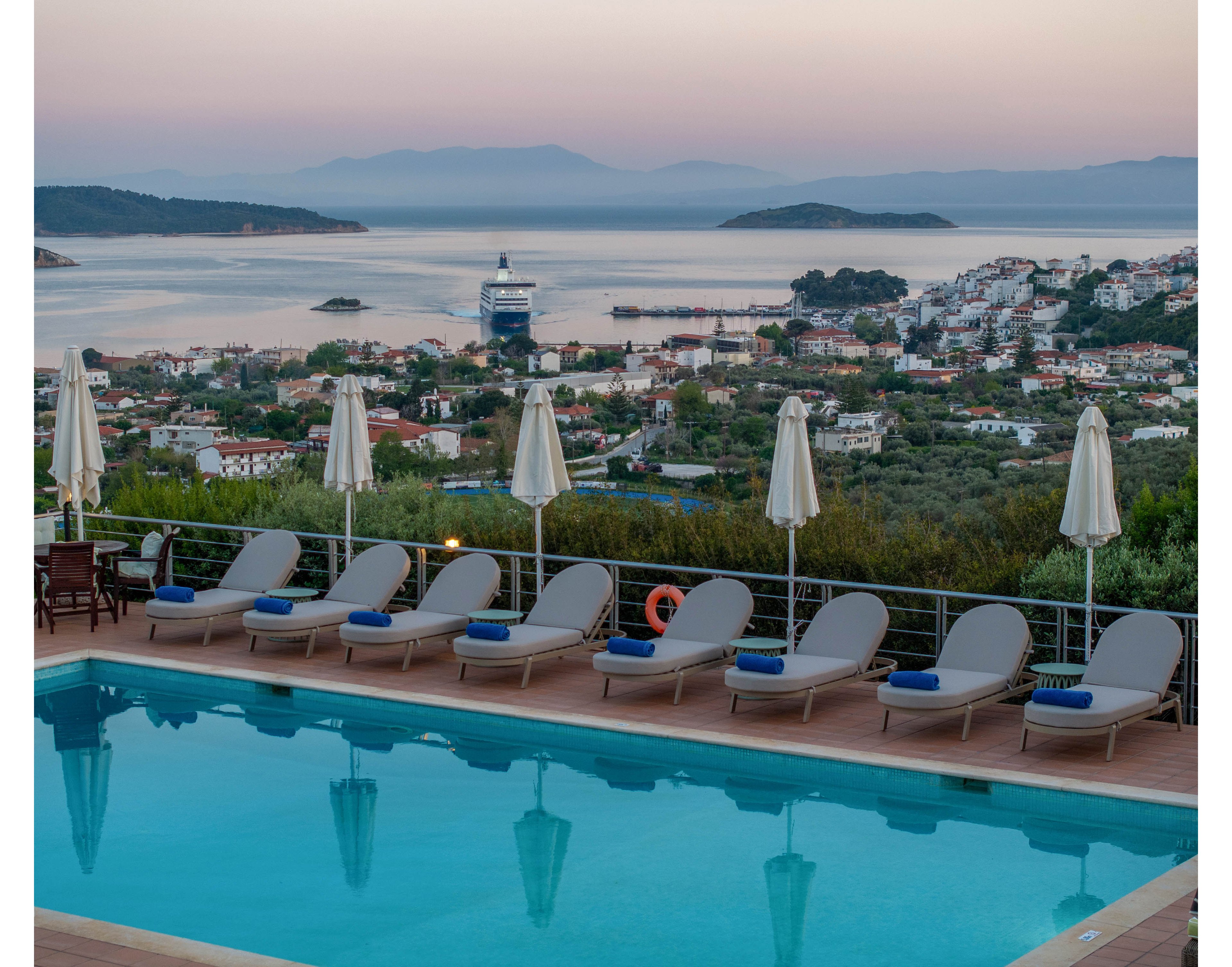 A serene hilltop swimming pool with lounge chairs and closed umbrellas overlooks a coastal town at dusk. Calm blue water in the pool reflects the soft pastel colors of the evening sky. In the distance, a large cruise ship sits in the bay surrounded by small islands and rolling hills. The scene conveys a peaceful vacation atmosphere with a focus on relaxation and scenic ocean views.