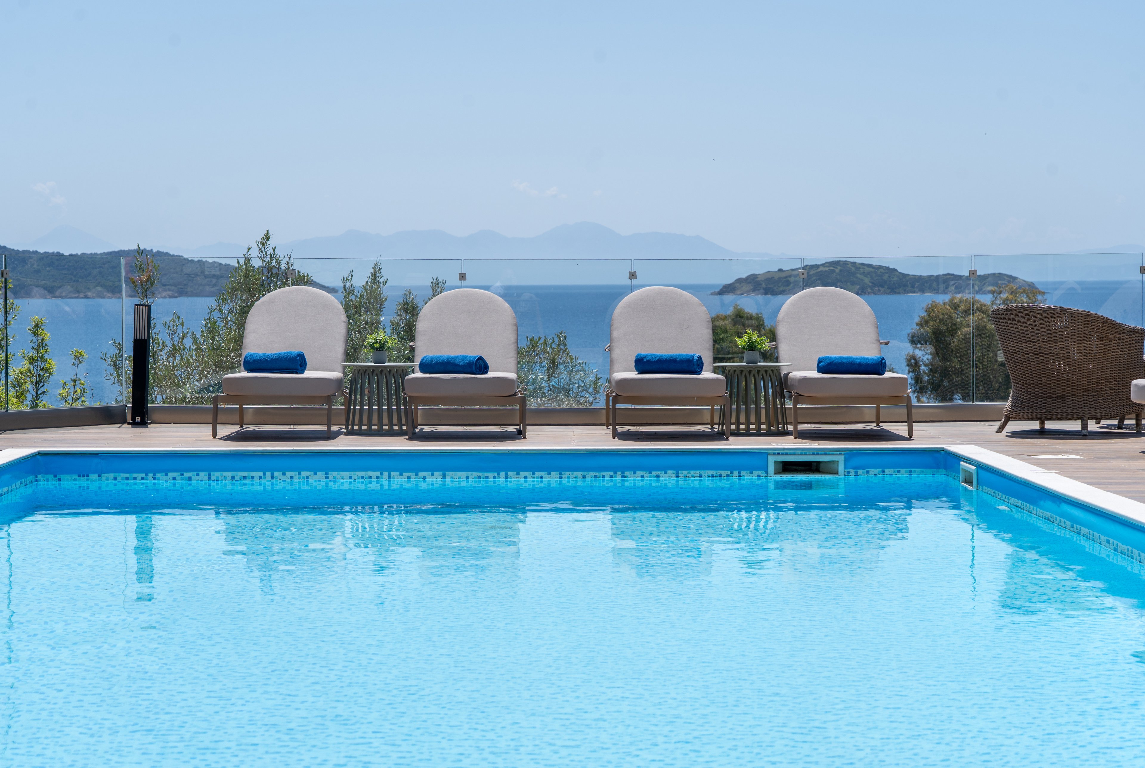 A row of cushioned sun loungers with blue towels faces a clear blue swimming pool. Beyond the glass railing, a calm sea and distant islands create a serene coastal backdrop. Wicker furniture in the foreground adds a touch of resort comfort and relaxation. The bright daylight and clear sky emphasize a peaceful vacation atmosphere.
