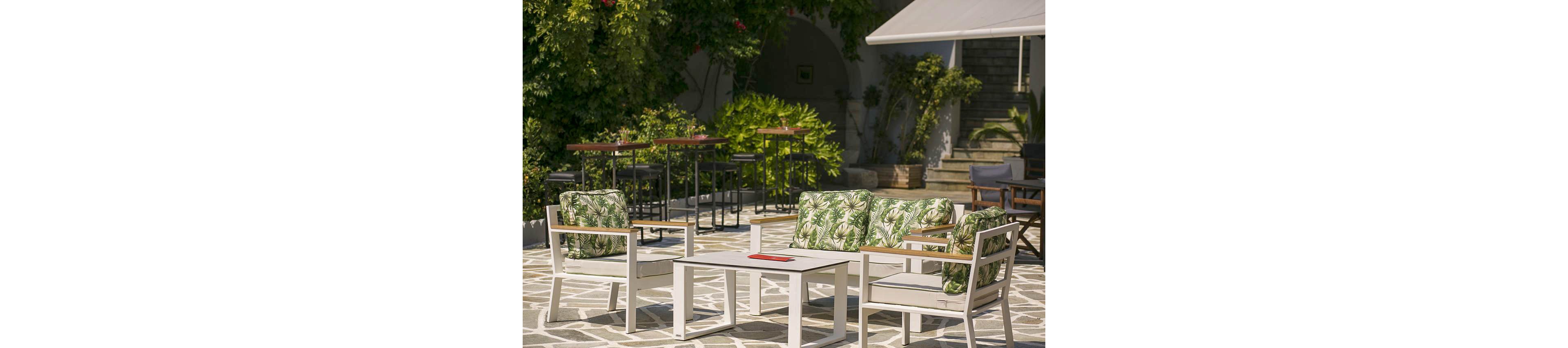 A bright outdoor patio features a white lounge seating set with green leaf-patterned cushions around a low coffee table. The area is paved with light stone tiles and surrounded by lush greenery and trees. In the background, tall bar tables and stools line the shaded edge of the courtyard. Sunlight and shadows create a relaxed, summery atmosphere ideal for socializing or relaxing.