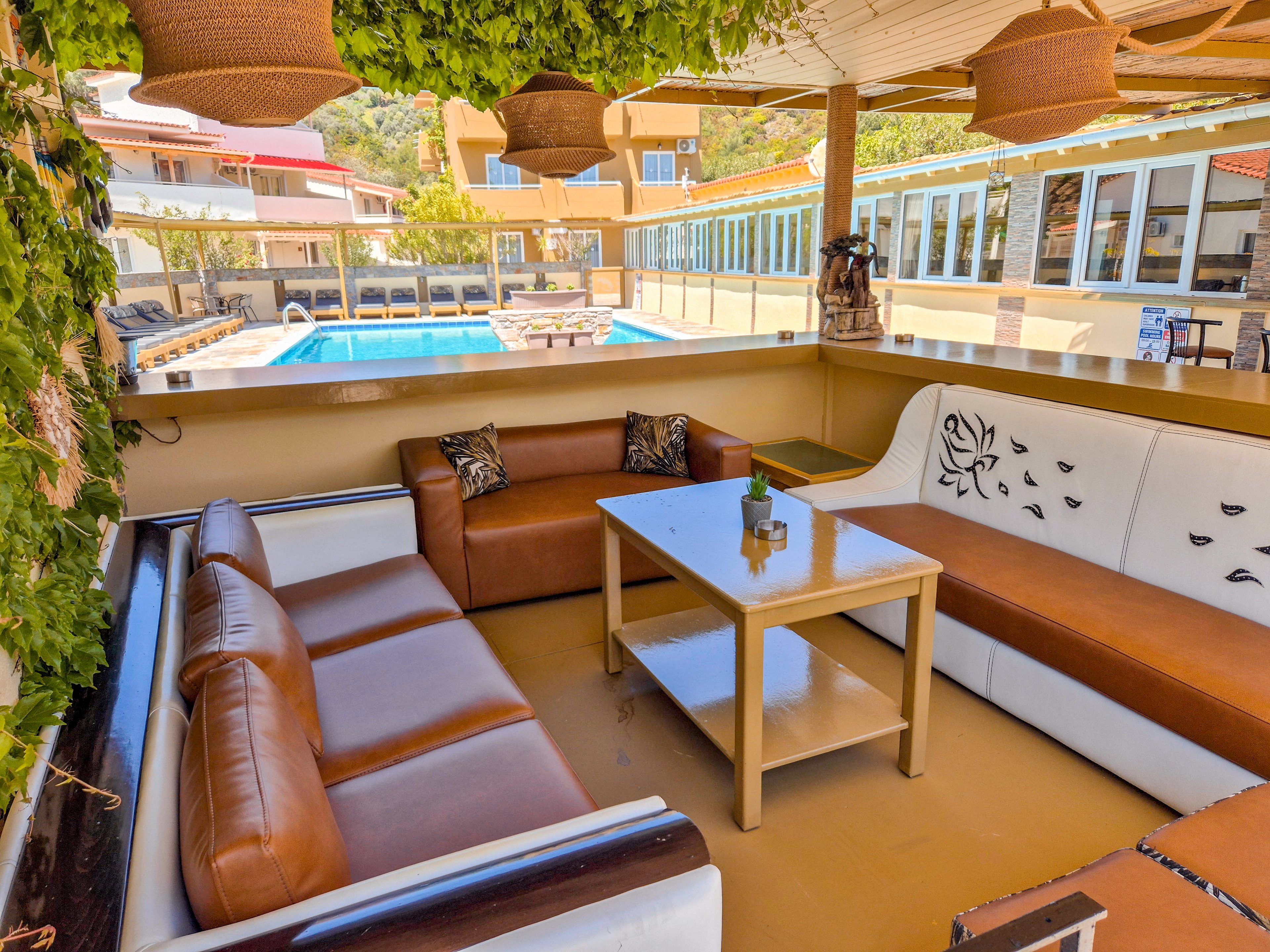 An outdoor lounge area features brown and white cushioned sofas arranged around a rectangular coffee table. The space is shaded by a pergola with hanging wicker lamps and green vines overhead. In the background, a clear blue swimming pool is surrounded by buildings with large windows and colorful roofs. The scene suggests a relaxed resort or hotel terrace designed for socializing and leisure.