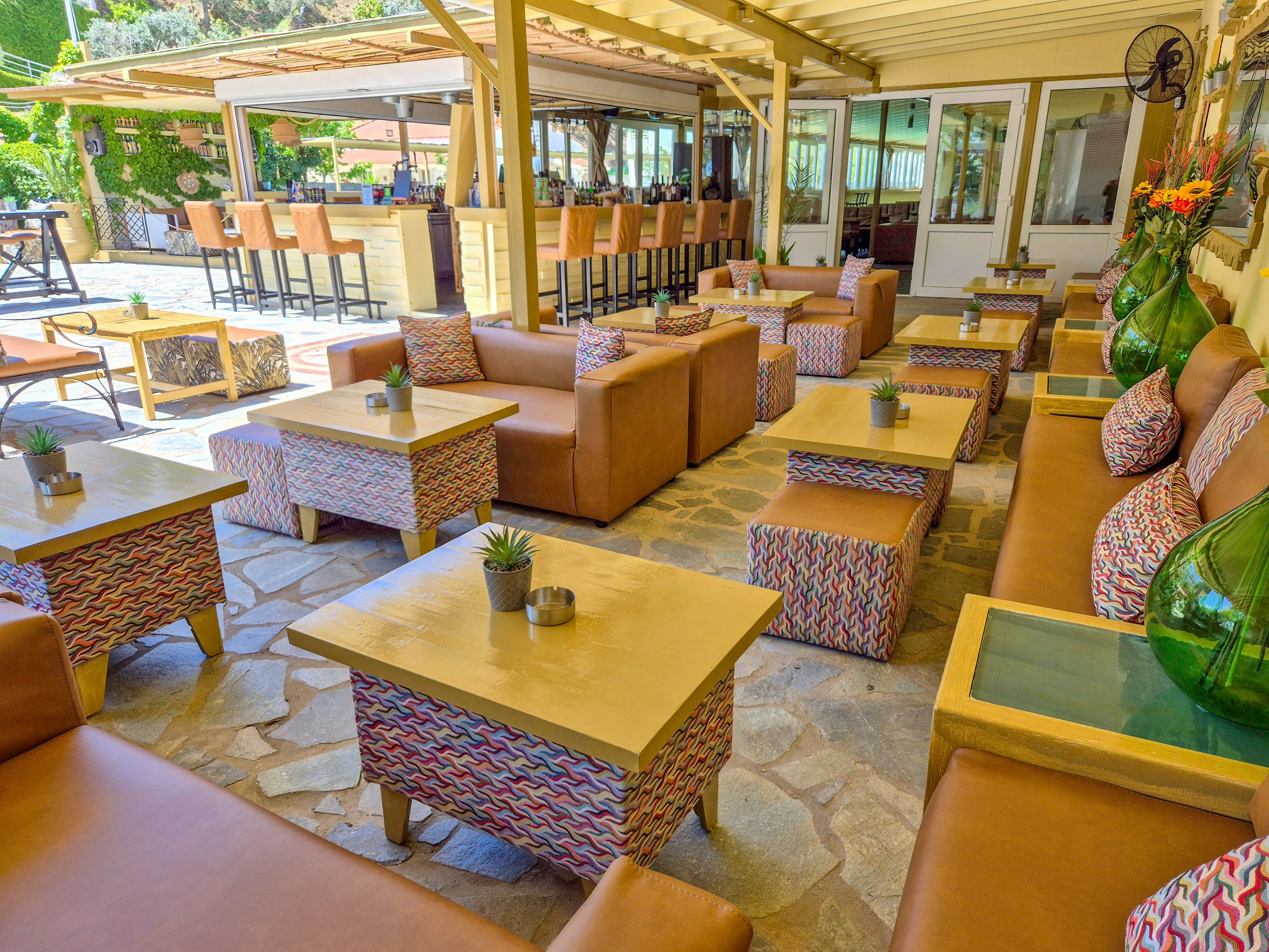 An outdoor lounge area features tan leather sofas and patterned cushioned stools arranged around low wooden tables. The space opens onto a bar with high stools under a covered pergola roof. Small potted plants and candles decorate the tabletops, adding a relaxed and inviting touch. Large glass vases with flowers and greenery enhance the casual resort atmosphere.