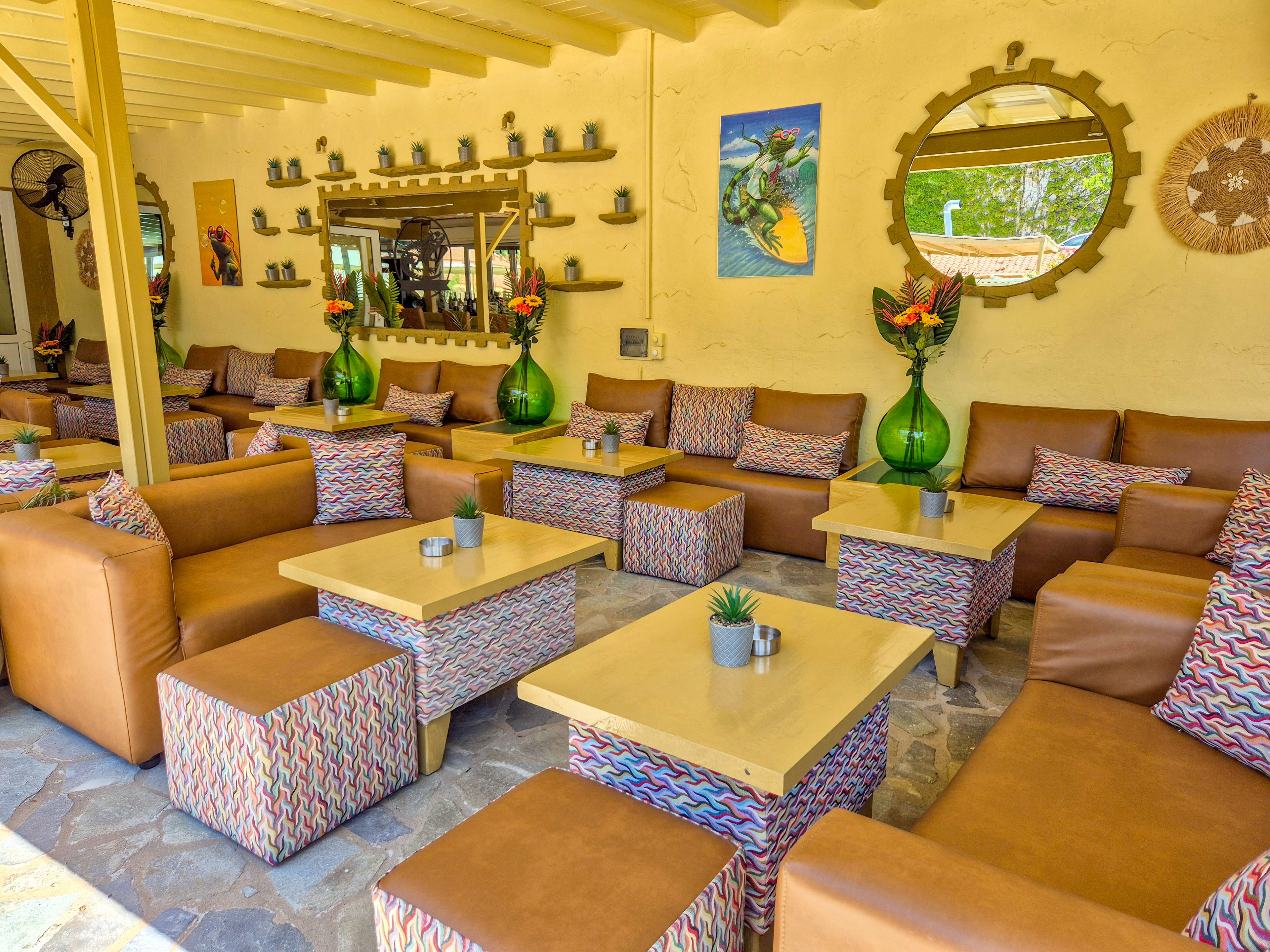 A stylish outdoor lounge area features multiple tan leather sofas and matching cube ottomans arranged around light wooden tables. Patterned fabric on the ottomans and cushions adds vibrant color to the relaxed seating layout. Large green glass vases with tropical flower arrangements decorate the tables, while small potted succulents provide additional accents. The background wall is adorned with mirrors, shelves of small pots, and colorful artwork, creating a cozy and inviting social space.