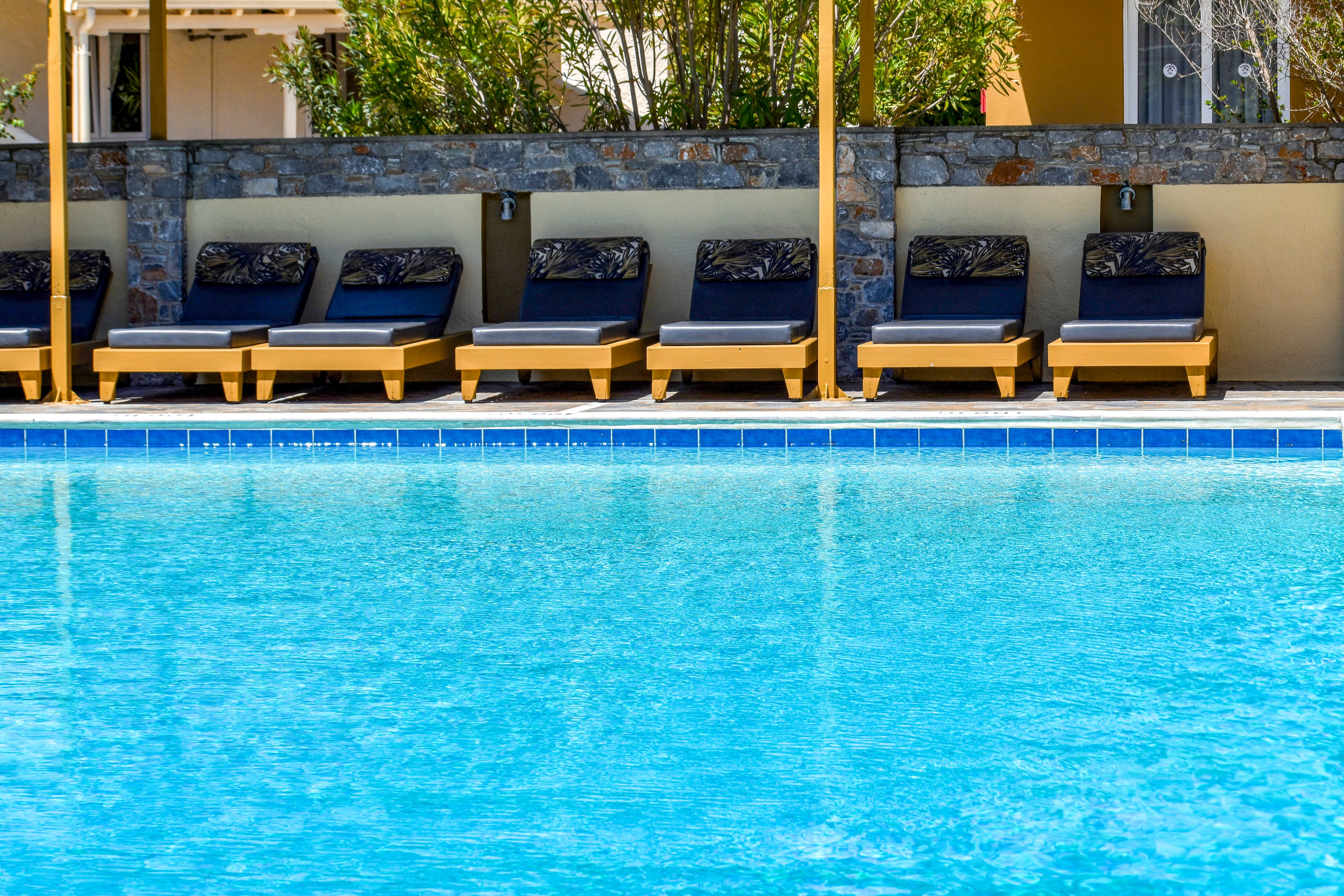 A clear blue swimming pool dominates the foreground, reflecting bright daylight. In the background, a row of cushioned sun loungers sits neatly aligned along the poolside. Stone walls and greenery provide a calm, private backdrop. The scene suggests a quiet resort or hotel area ready for guests to relax by the water.