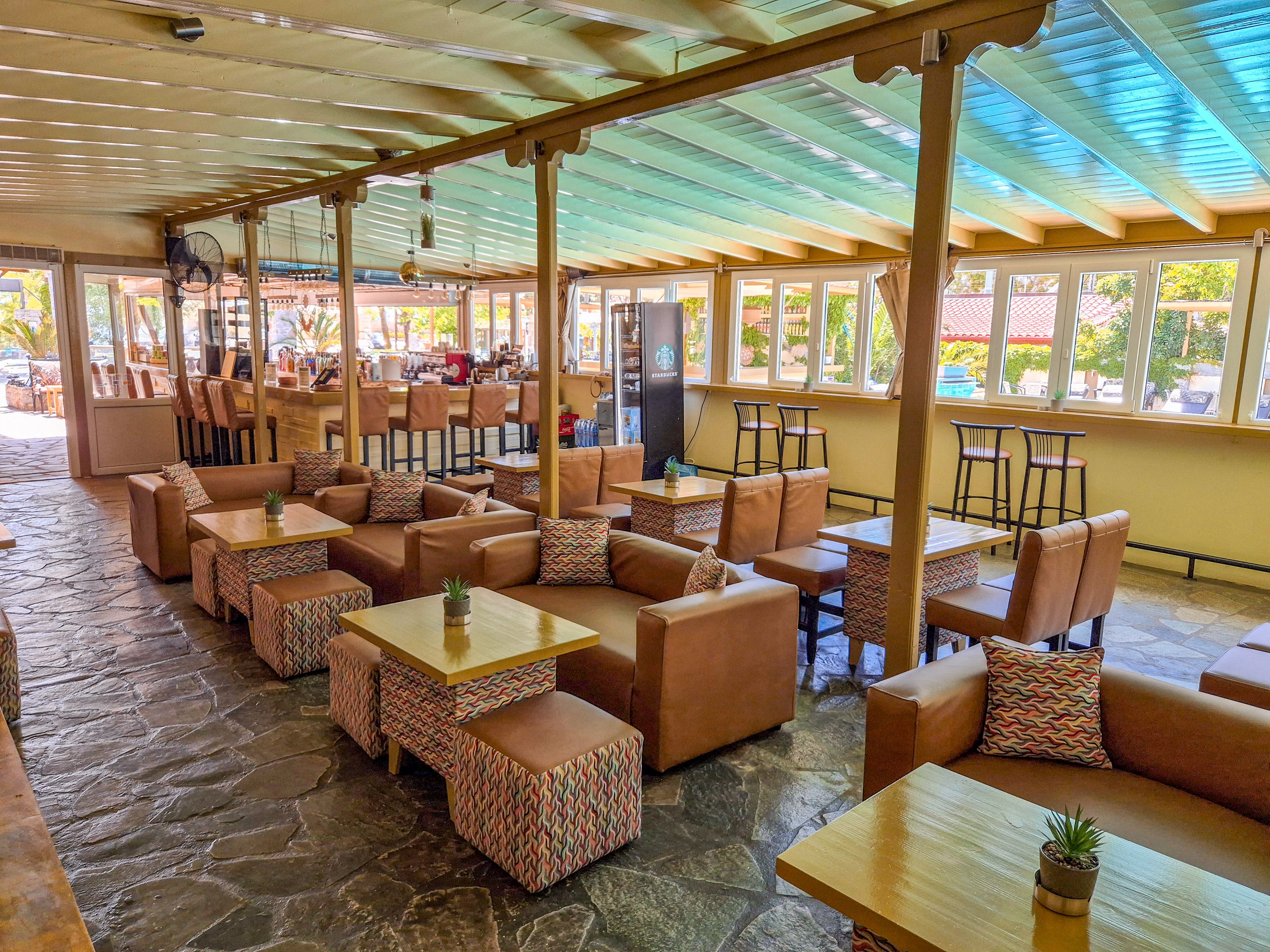 Spacious indoor lounge bar featuring rows of cushioned armchairs and low wooden tables. The setting includes a stone floor, high bar stools near the windows, and a long bar counter in the background. Natural light streams through large windows and a translucent ceiling, creating a bright and airy atmosphere. Colorful patterned cushions and small potted plants on the tables add decorative accents to the relaxed social space.