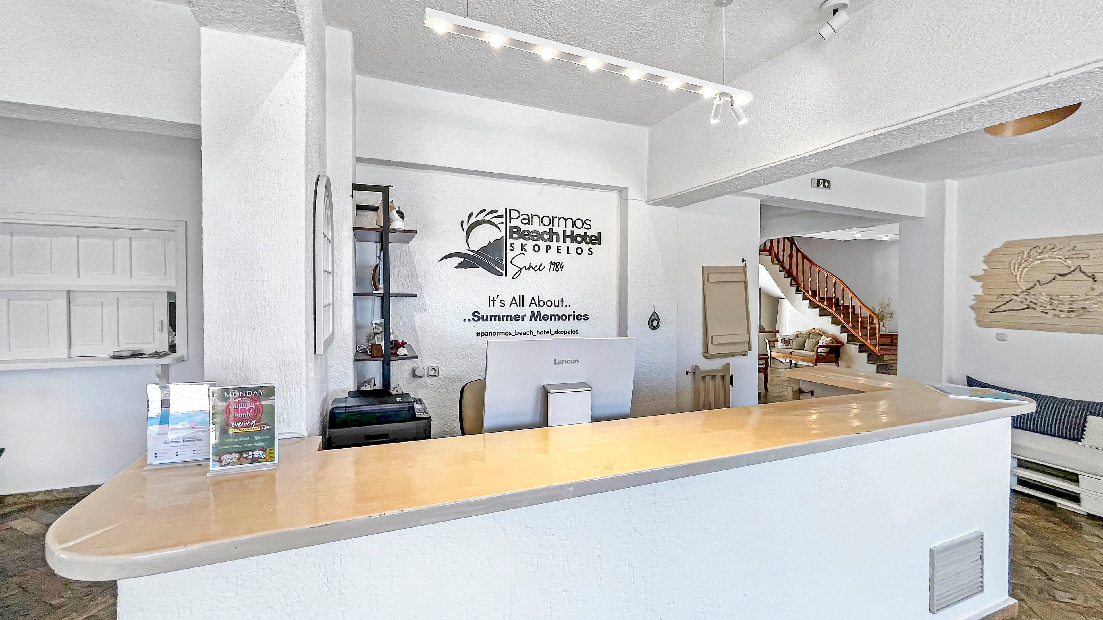 A bright hotel reception area features a long, light-colored front desk with brochures and a payment terminal on top. Behind the counter, a wall sign reads "Panormos Beach Hotel" and includes the phrase "It’s All About Summer Memories" along with decorative graphics. The space is painted white with modern lighting, open shelving, and a doorway leading to a staircase. A seating area with cushions and wall art is visible to the right, enhancing the welcoming atmosphere.