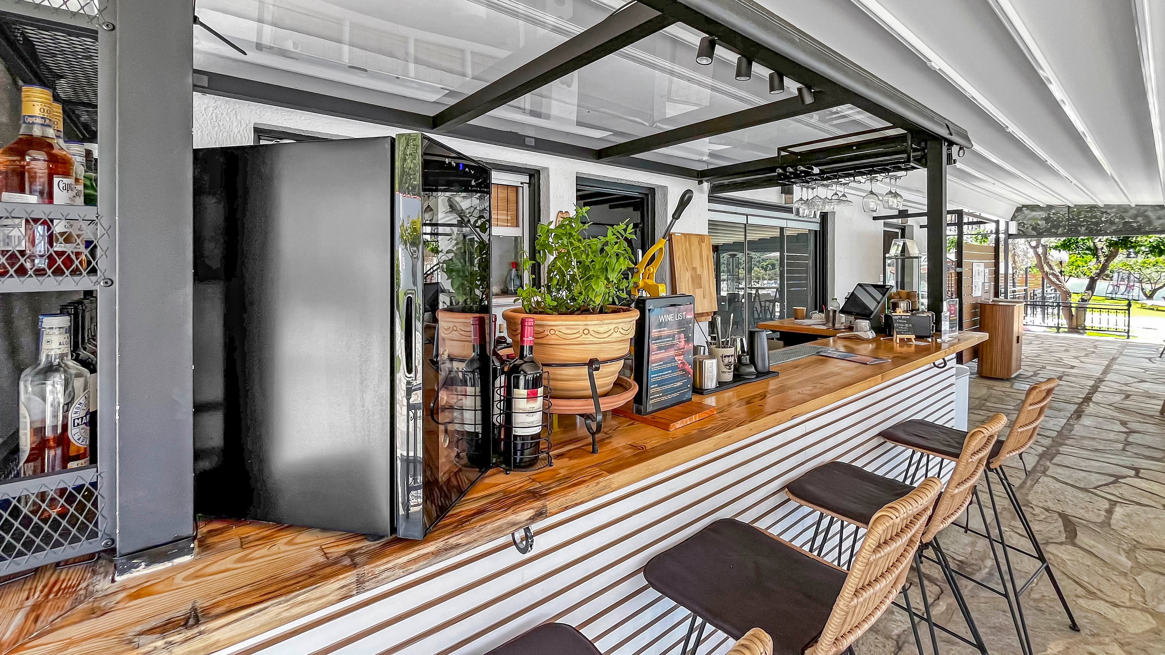 An outdoor bar counter features a wooden surface lined with wicker bar stools. Bottles, glassware, and a potted plant sit on the counter beside a black refrigerator. Shelving with various spirits is visible on the left, while large windows and doors open to the patio area. The bright, covered terrace and stone flooring create a relaxed, airy atmosphere suitable for social gatherings.