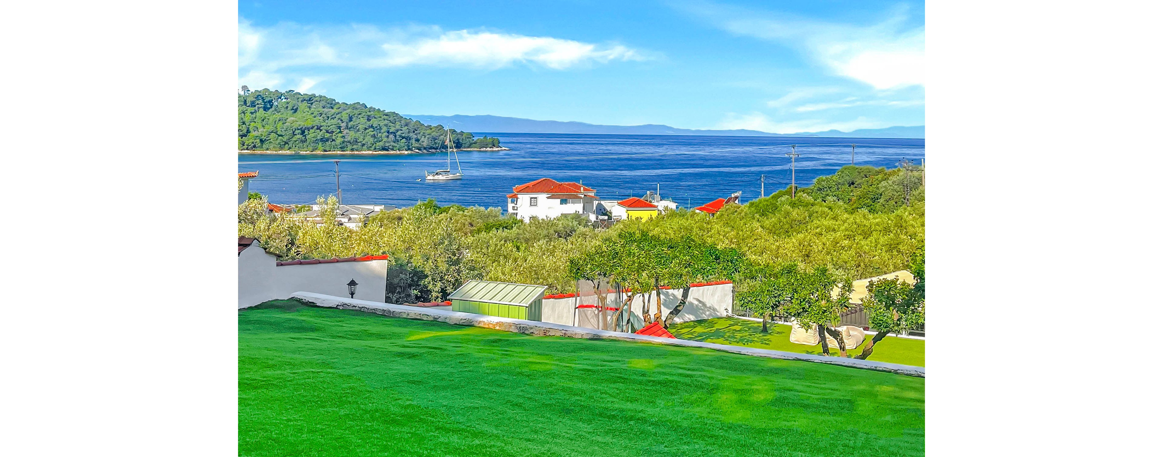 A vibrant coastal landscape overlooks a calm blue sea with sailboats in the distance. White houses with red roofs sit among lush green trees on a gentle hillside. In the foreground, a bright green lawn and garden structures lead the eye toward the shoreline. The scene is bathed in clear daylight under a blue sky with soft clouds, creating a peaceful vacation atmosphere.